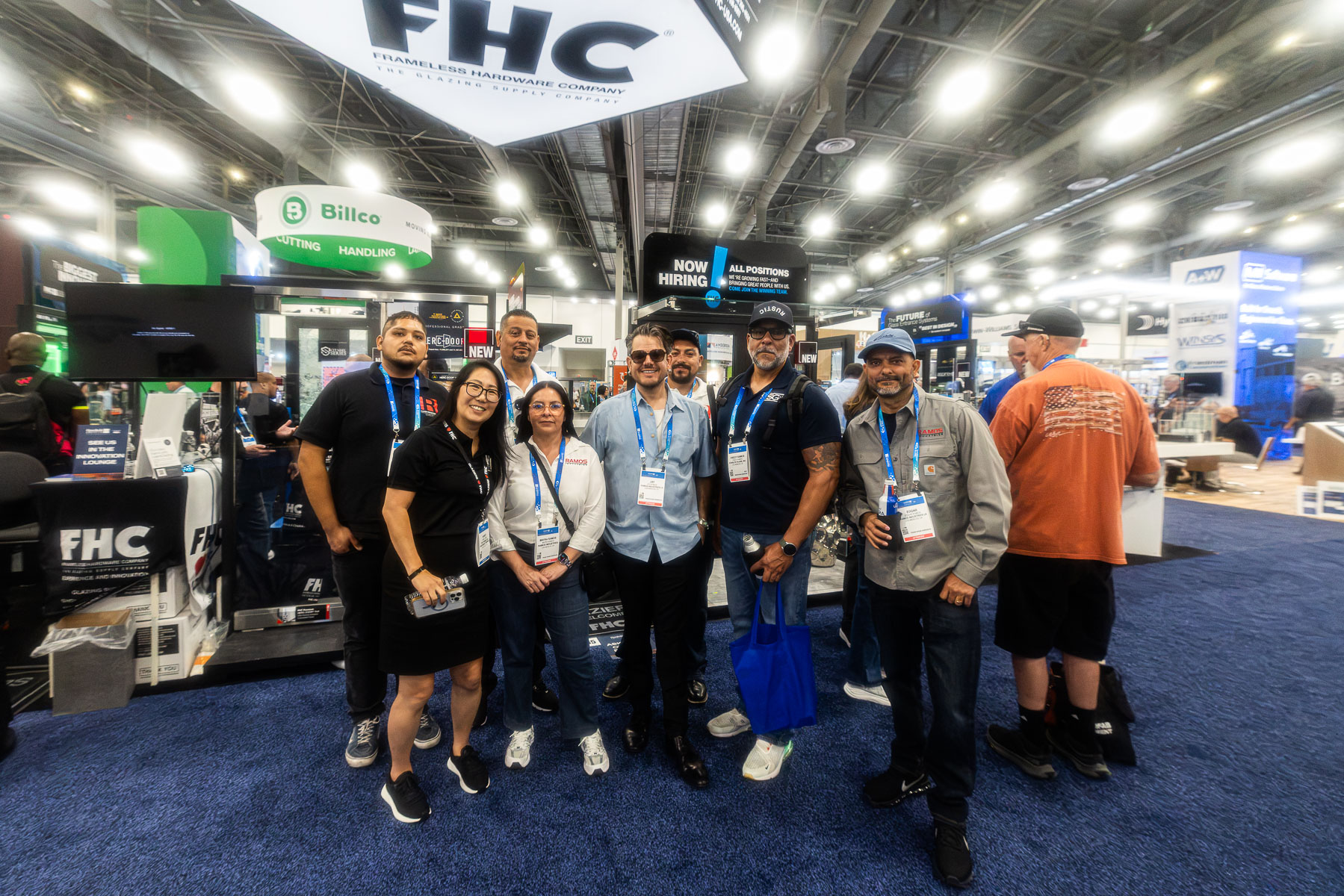 A group of people stands smiling at the FHC (Frameless Hardware Company) booth on a blue carpeted exhibition floor.