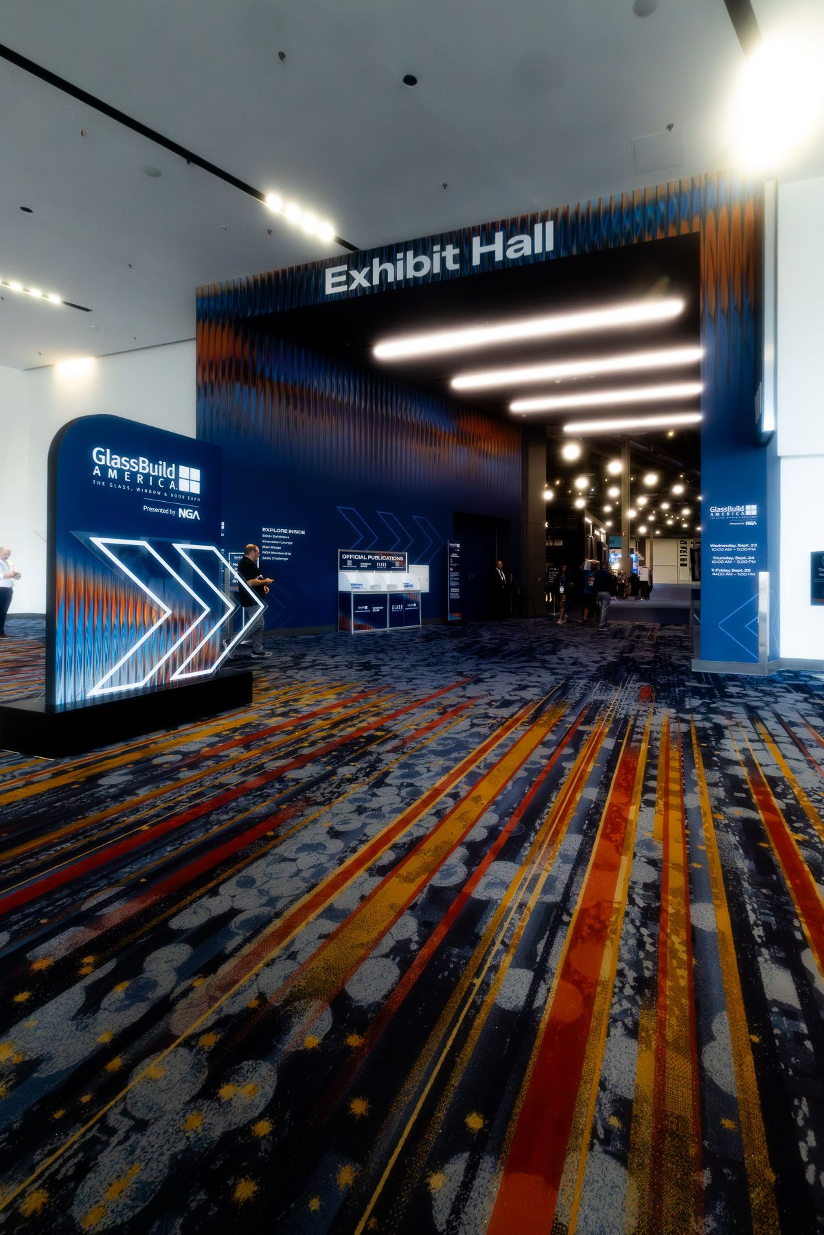 An illuminated entrance to an "Exhibit Hall" with GlassBuild America branding and directional signage on a patterned carpet.