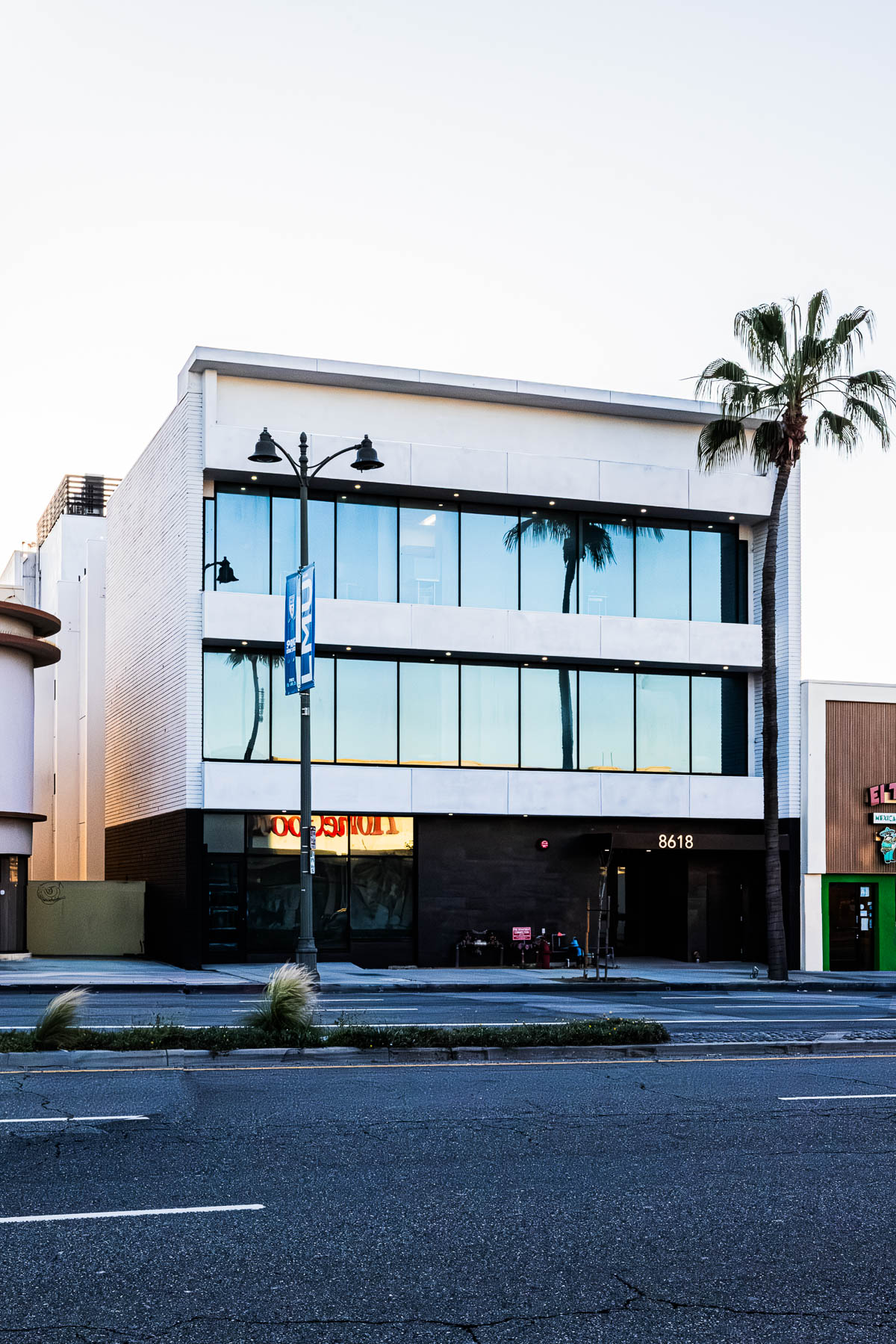 An exterior view of a modern three-story commercial building with large glass windows, dark ground floor panels, and a palm tree on the right.
