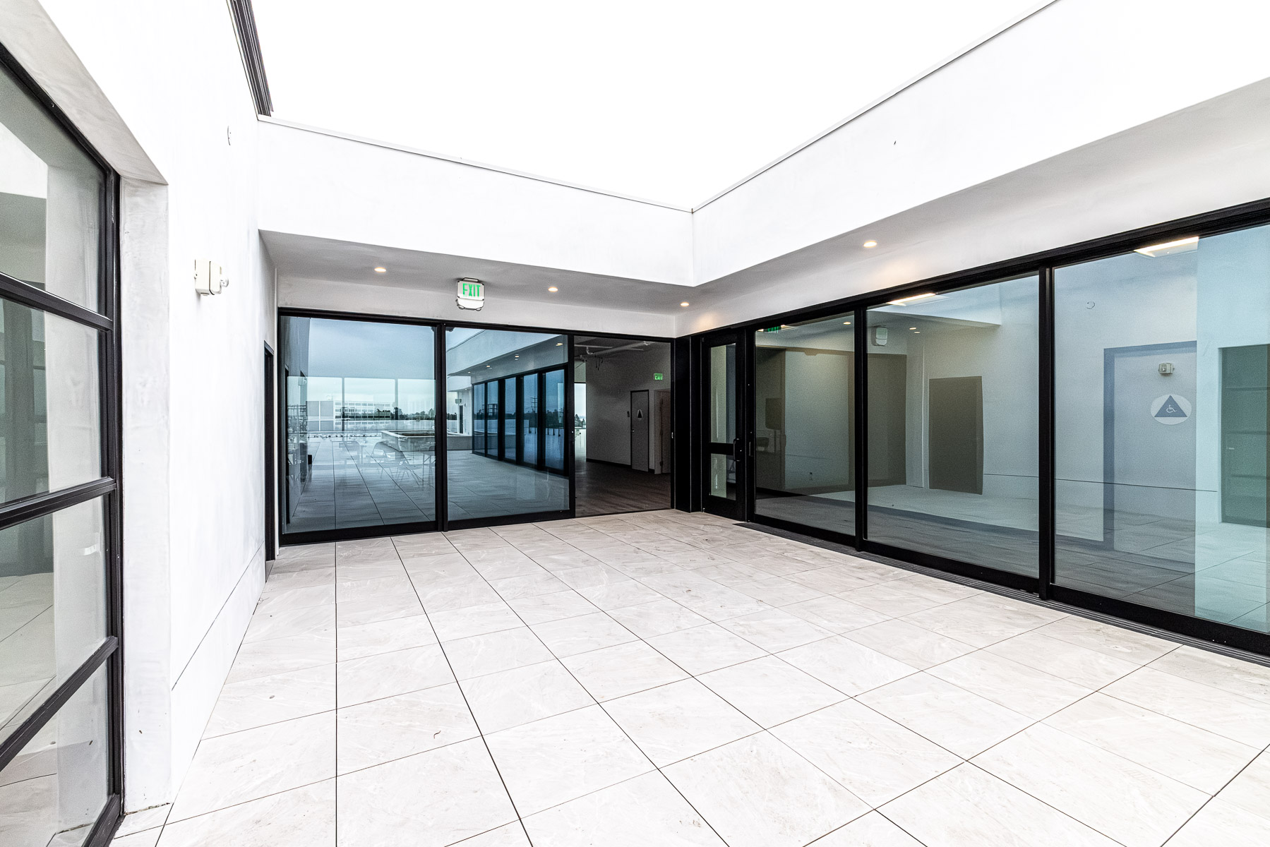 A bright, modern hallway with white tiled floors and large black-framed glass storefronts, including Arcadia multi-slider doors, offering views to an outdoor patio and interior rooms.