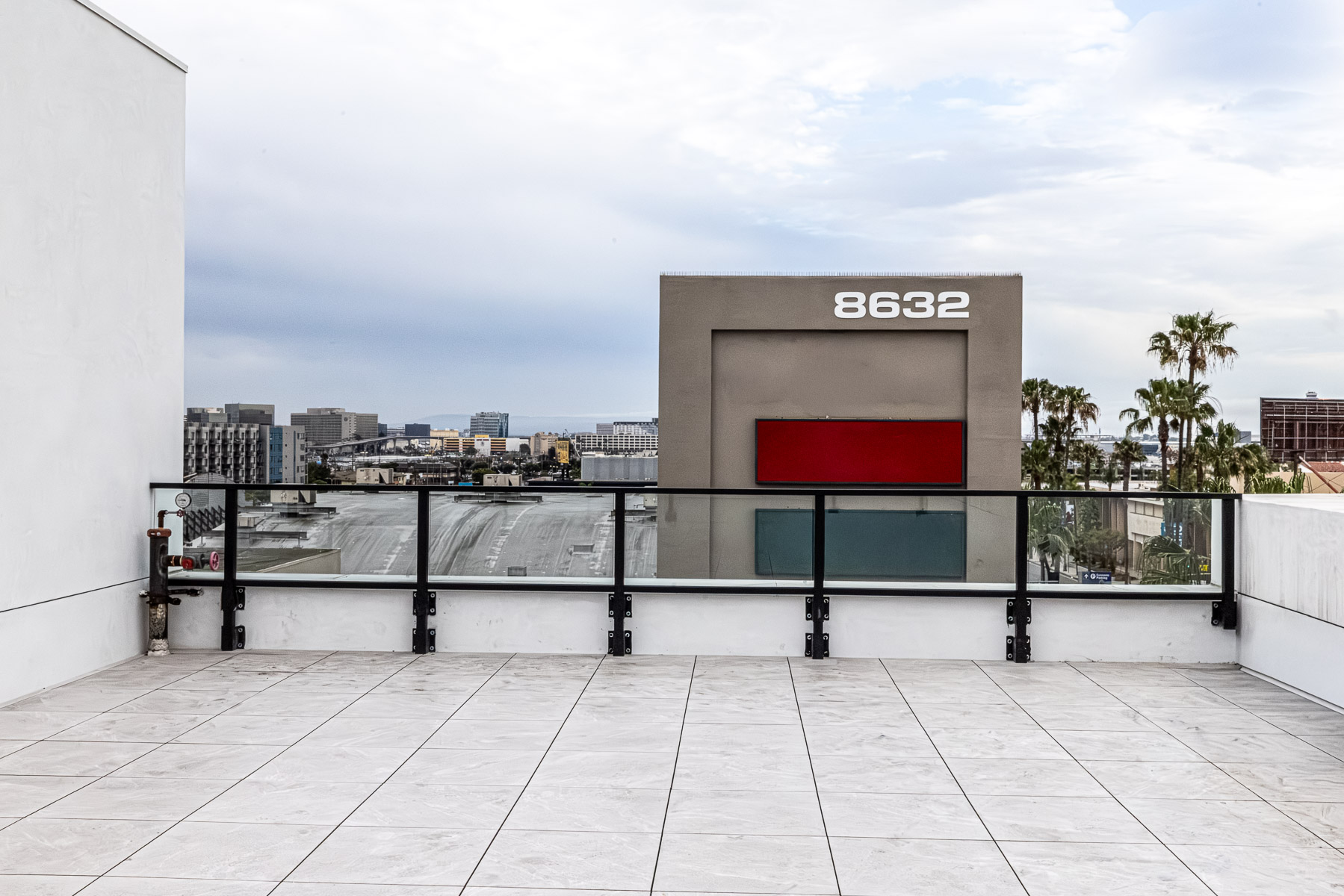 A rooftop patio with a glass and aluminum railing system overlooks a cityscape with a building displaying the address '8632'.