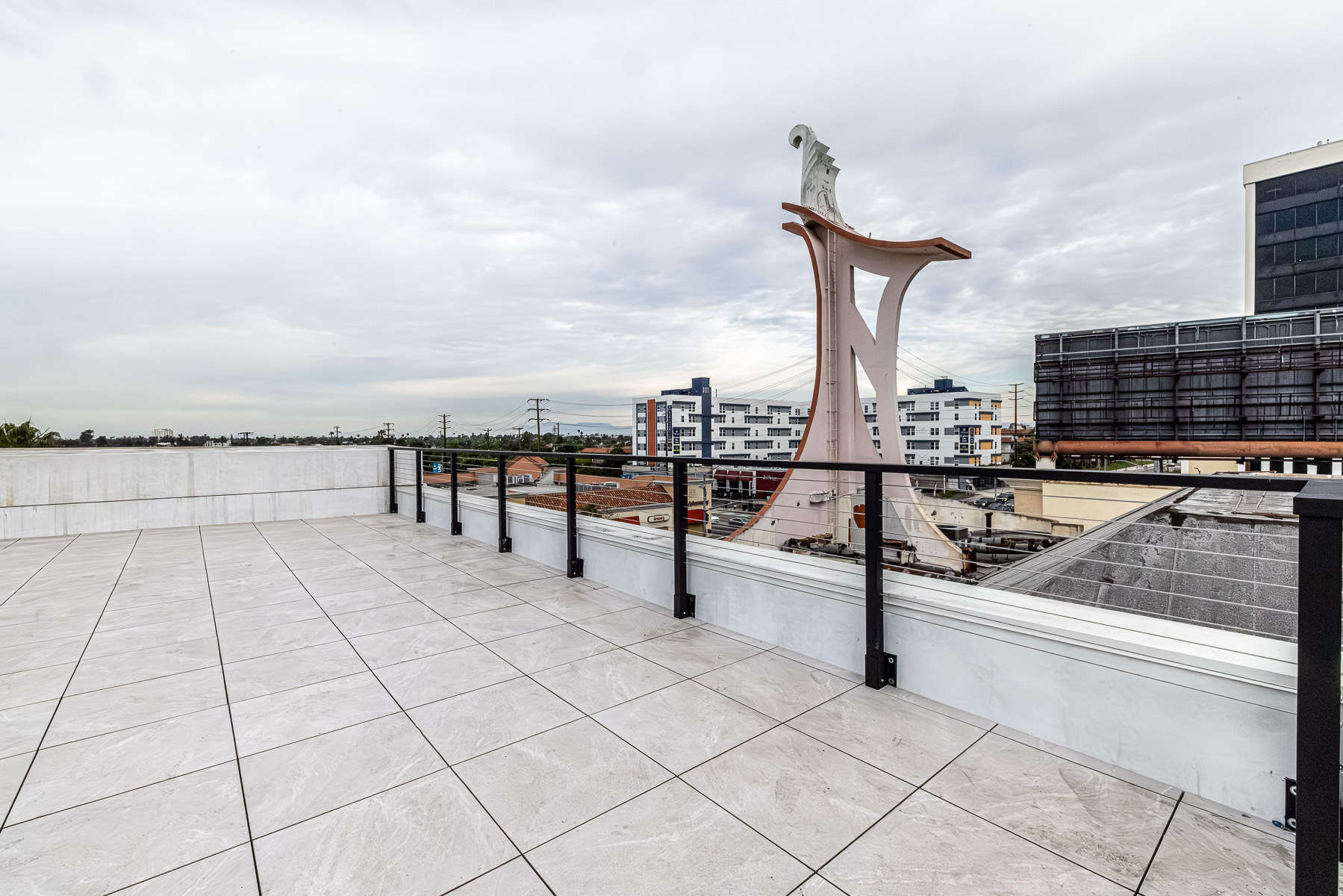 A rooftop patio with light-colored tiles and a black cable railing system overlooking a city skyline under an overcast sky.