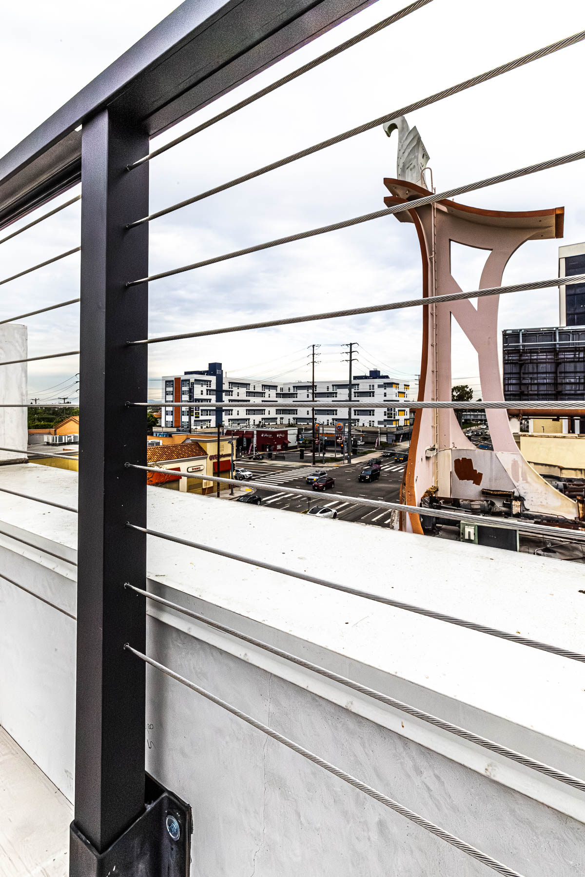 A close-up view of a dark metal post and horizontal stainless steel cables of a railing system, overlooking a city street with buildings and a large abstract sculpture under a cloudy sky.