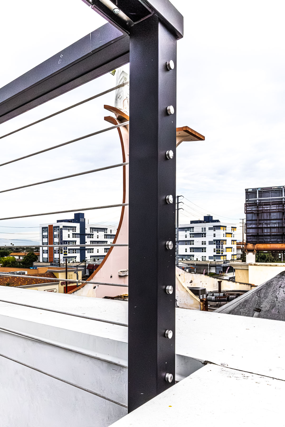 A close-up view of a dark metal cable railing system with horizontal steel cables, overlooking an urban landscape with buildings under an overcast sky.