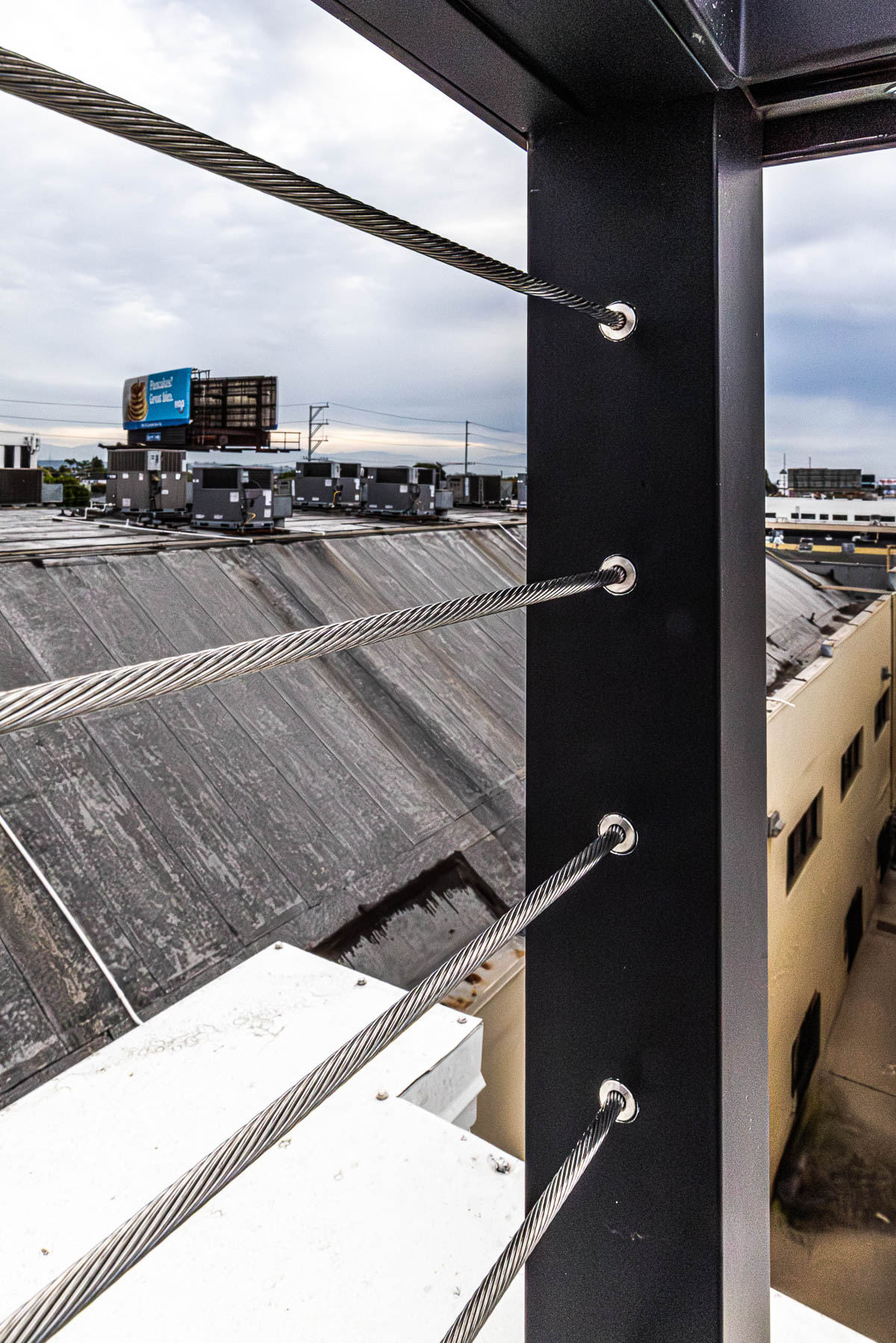 A close-up view of a black aluminum railing post with multiple stainless steel cables running horizontally through it, overlooking a rooftop with HVAC units and a billboard.