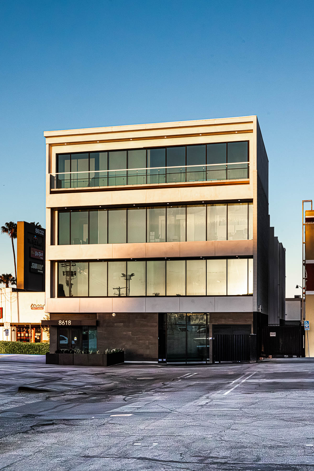 A modern three-story commercial building with extensive glass storefronts and a top-floor balcony with glass and aluminum railings, viewed from a parking lot under a clear blue sky.