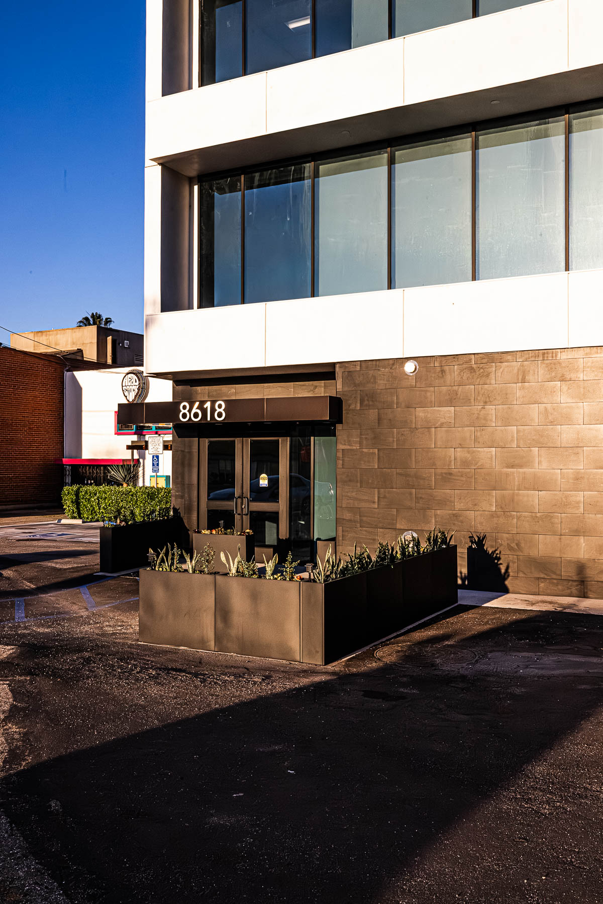 A modern building entrance with glass doors, dark tiled walls, white upper facade, and planters under a clear blue sky.