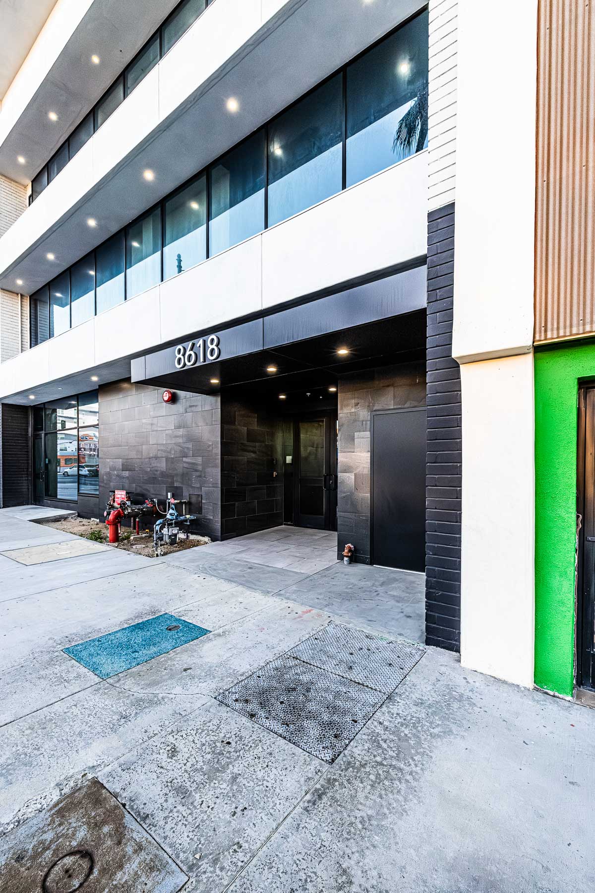 Exterior view of a modern building entrance with large glass storefront windows, dark tile cladding, and the address number 8618.