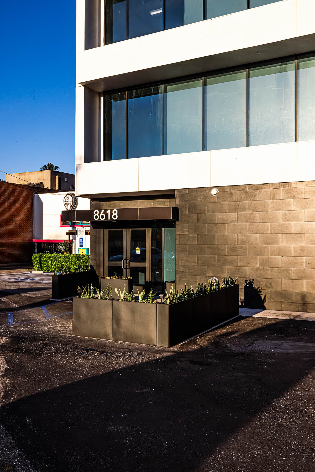 Exterior view of a modern commercial building entrance featuring glass storefront doors, dark wall panels, and planter boxes.