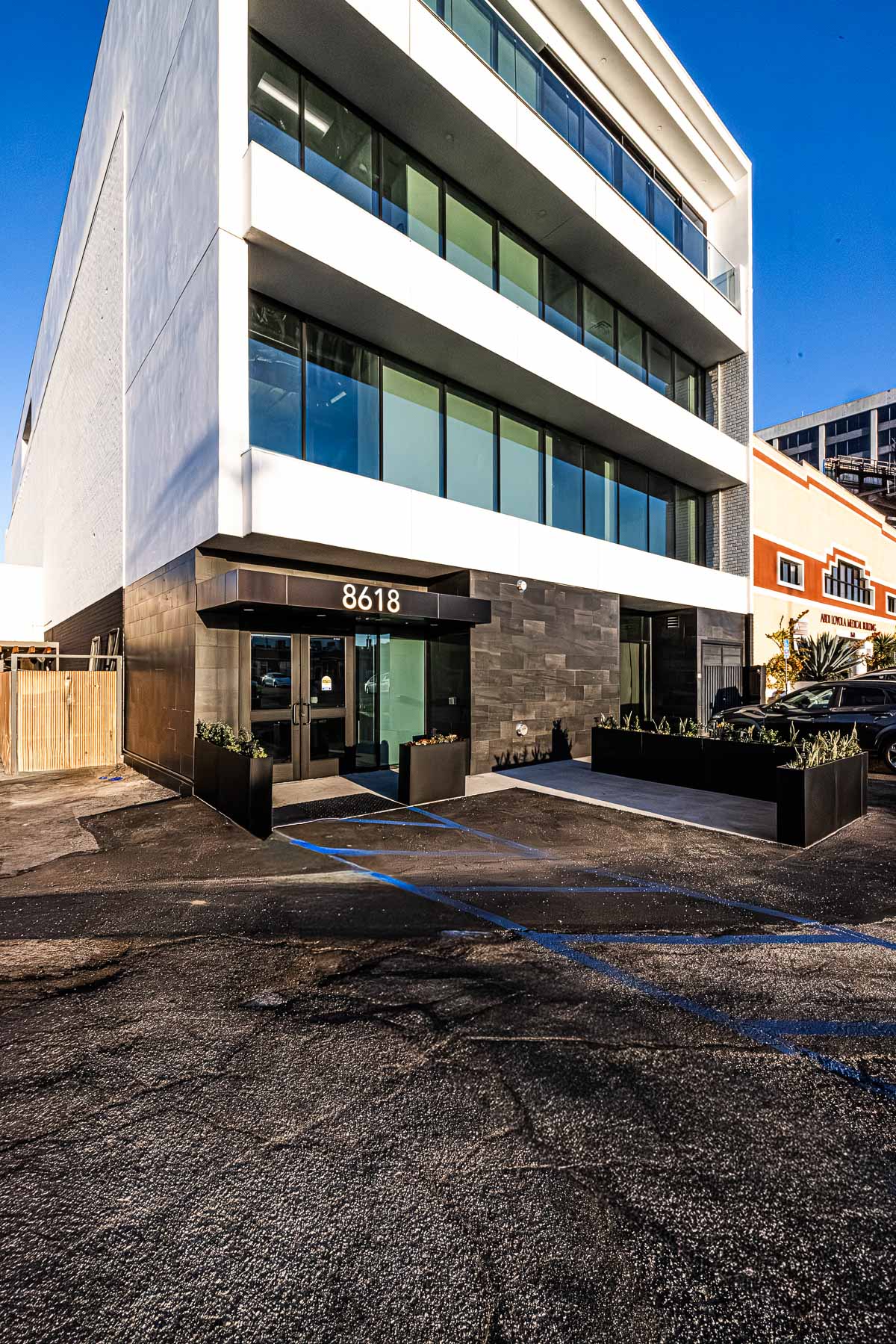 A modern multi-story building with a white upper facade and dark gray lower section, featuring large glass windows, an entrance with '8618' above it, and a parking lot.