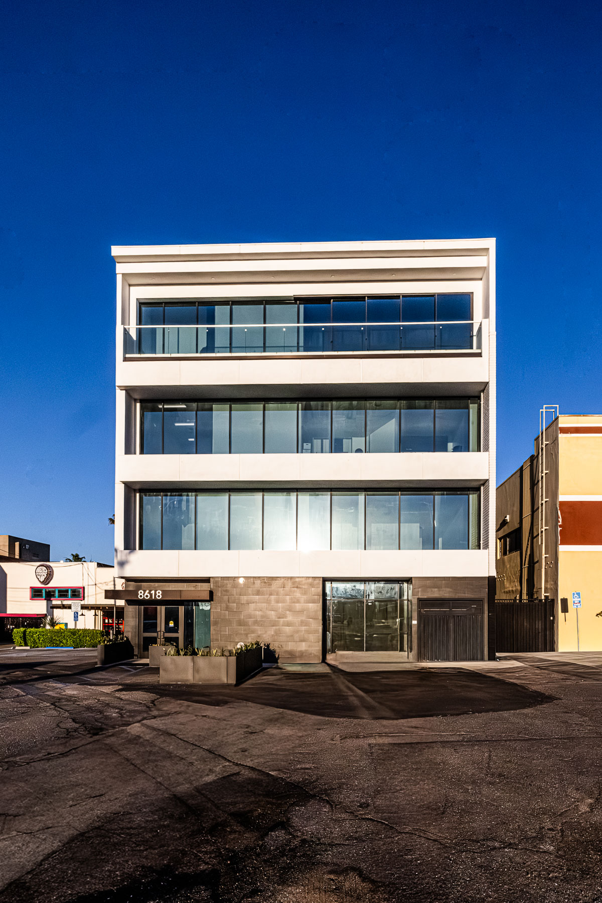 Exterior view of a modern, multi-story commercial building featuring a glass facade, storefront doors, and a balcony with a railing.