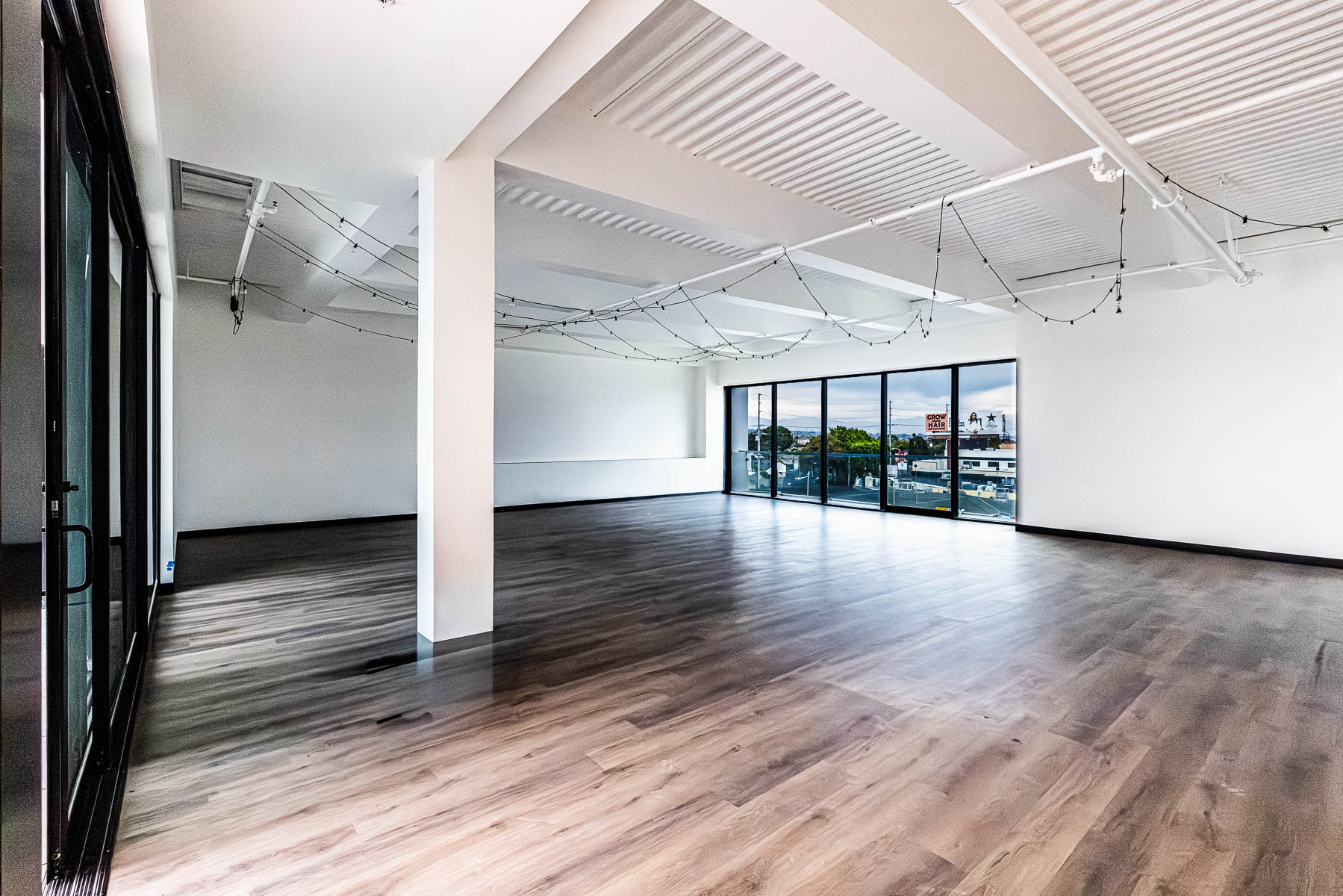 An empty, modern room with wood-look flooring, white walls, and large dark-framed glass doors and windows overlooking a city.