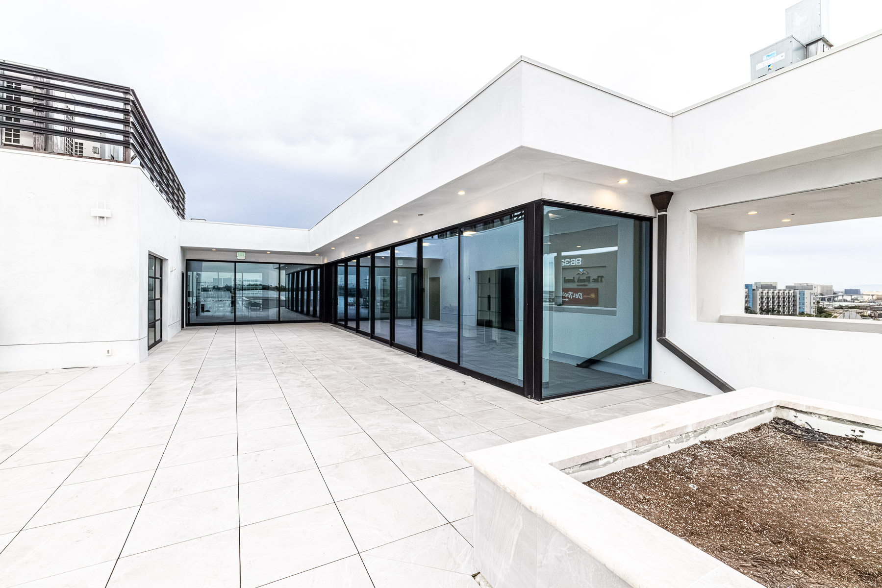 An exterior rooftop patio features a long stretch of modern glass storefront with dark frames, multi-sliding doors, and a light-tiled floor, with a cityscape visible in the background.