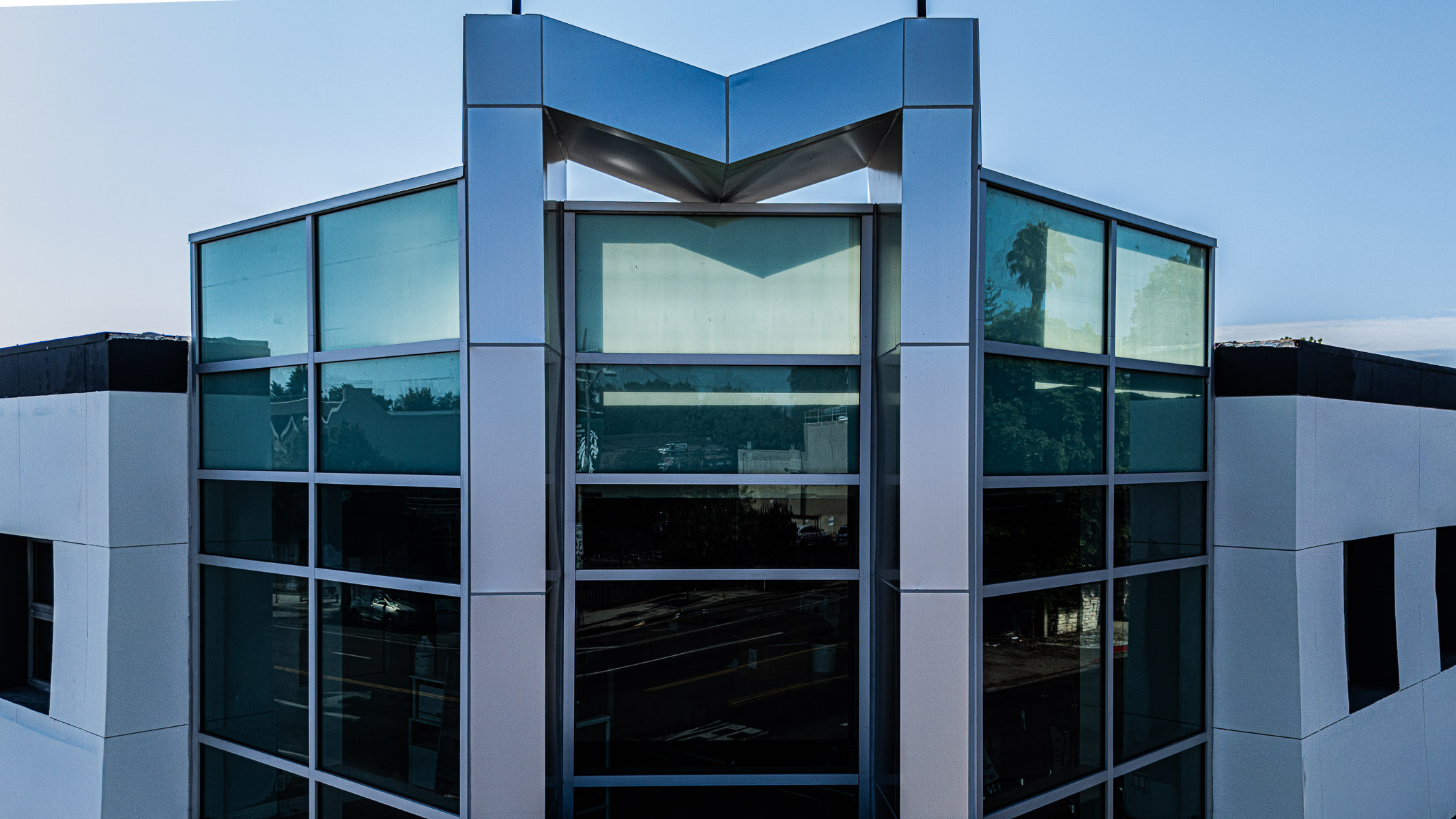 An exterior view of a modern commercial building's corner, featuring a multi-paneled glass curtainwall with a distinctive metallic 'V' shape at the top.