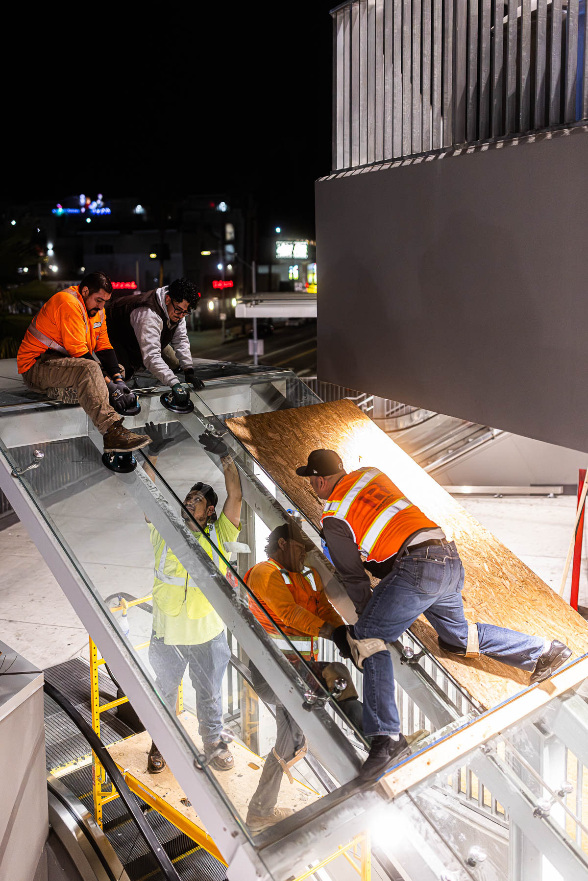 Four construction workers install a large clear glass panel on an exterior building entrance at night, with city lights in the background.