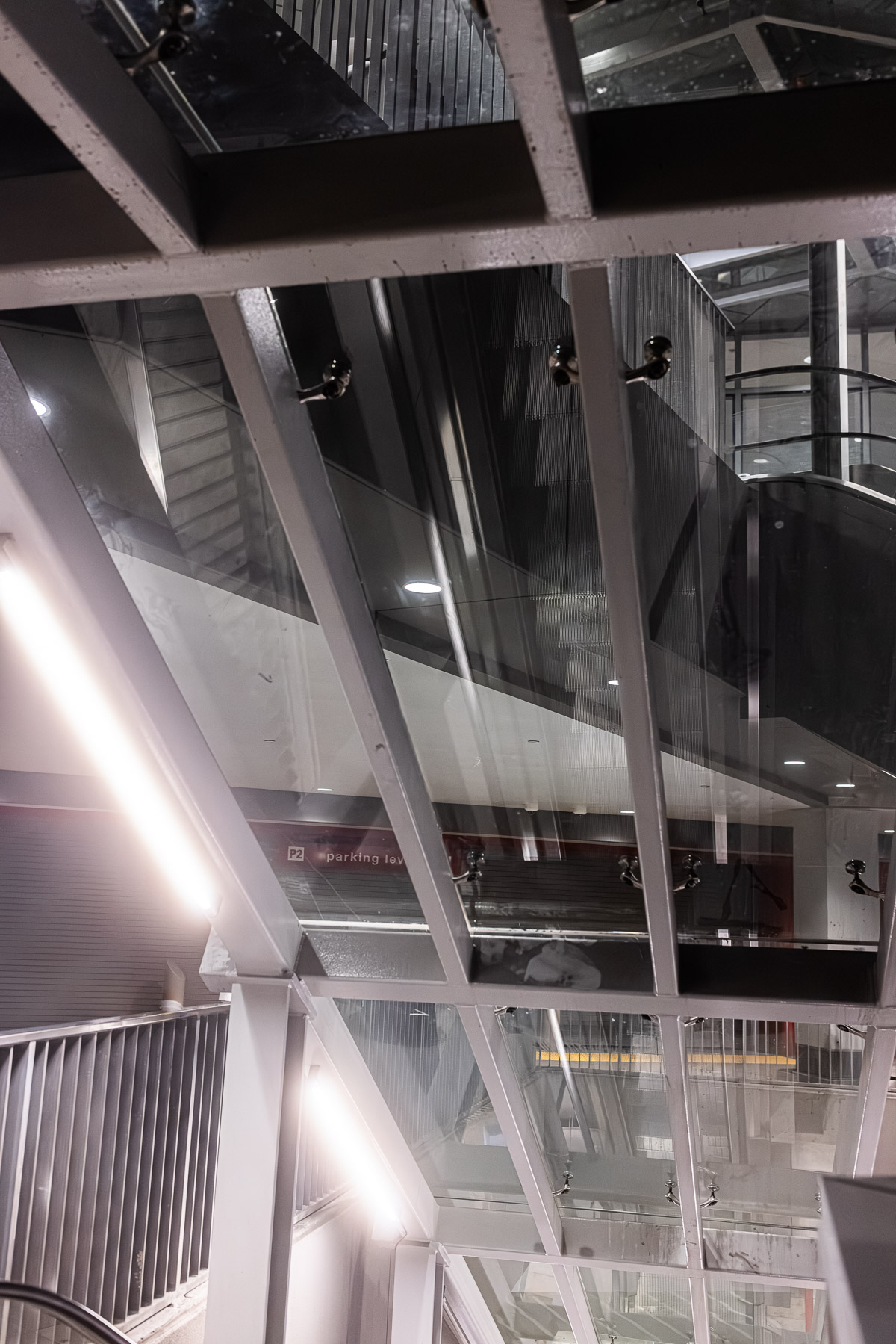A view looking up through a clear glass canopy supported by metal beams and spider fittings, revealing an escalator and interior building elements beyond.