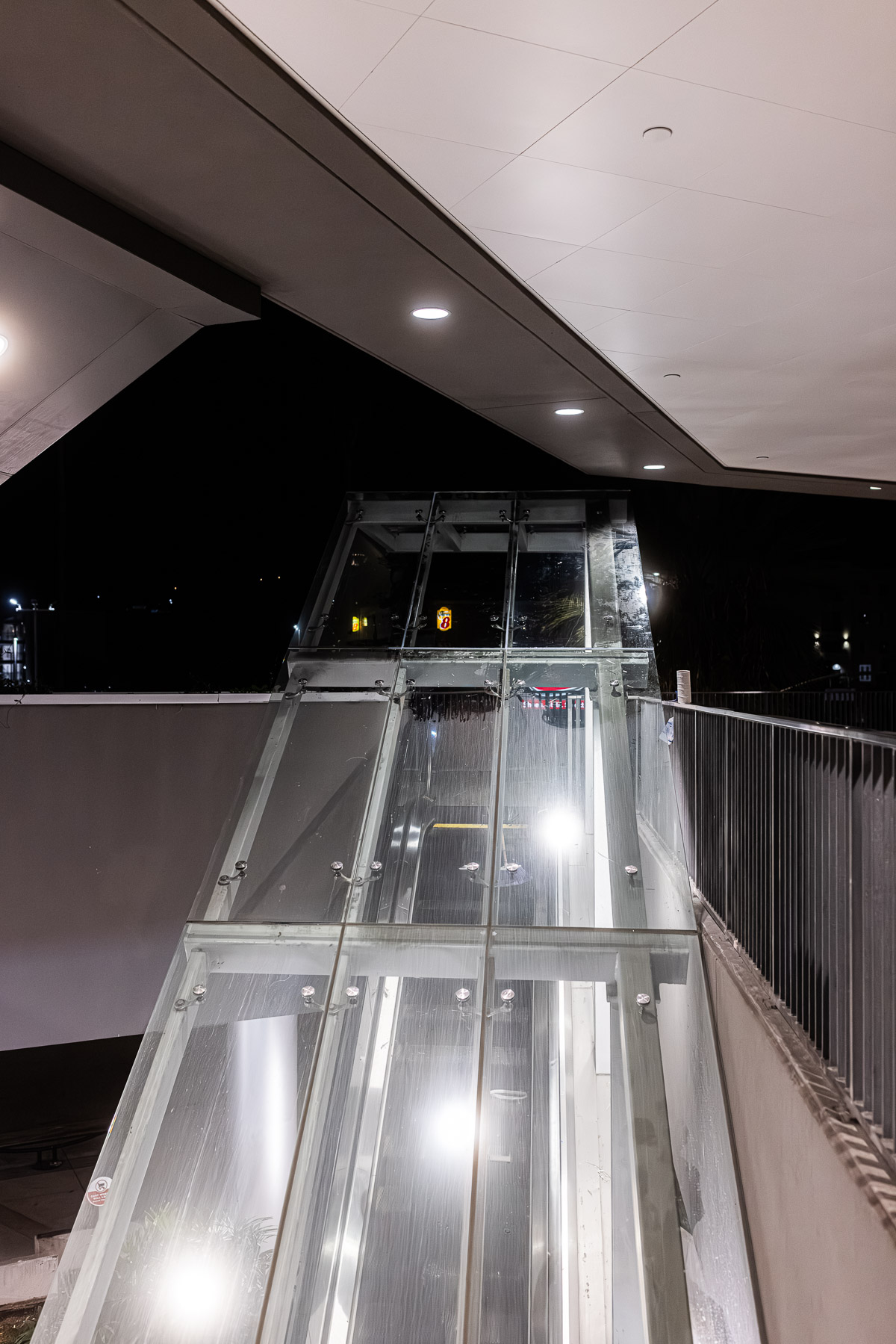An exterior night view of a modern glass canopy and escalator entrance, illuminated by overhead lights.