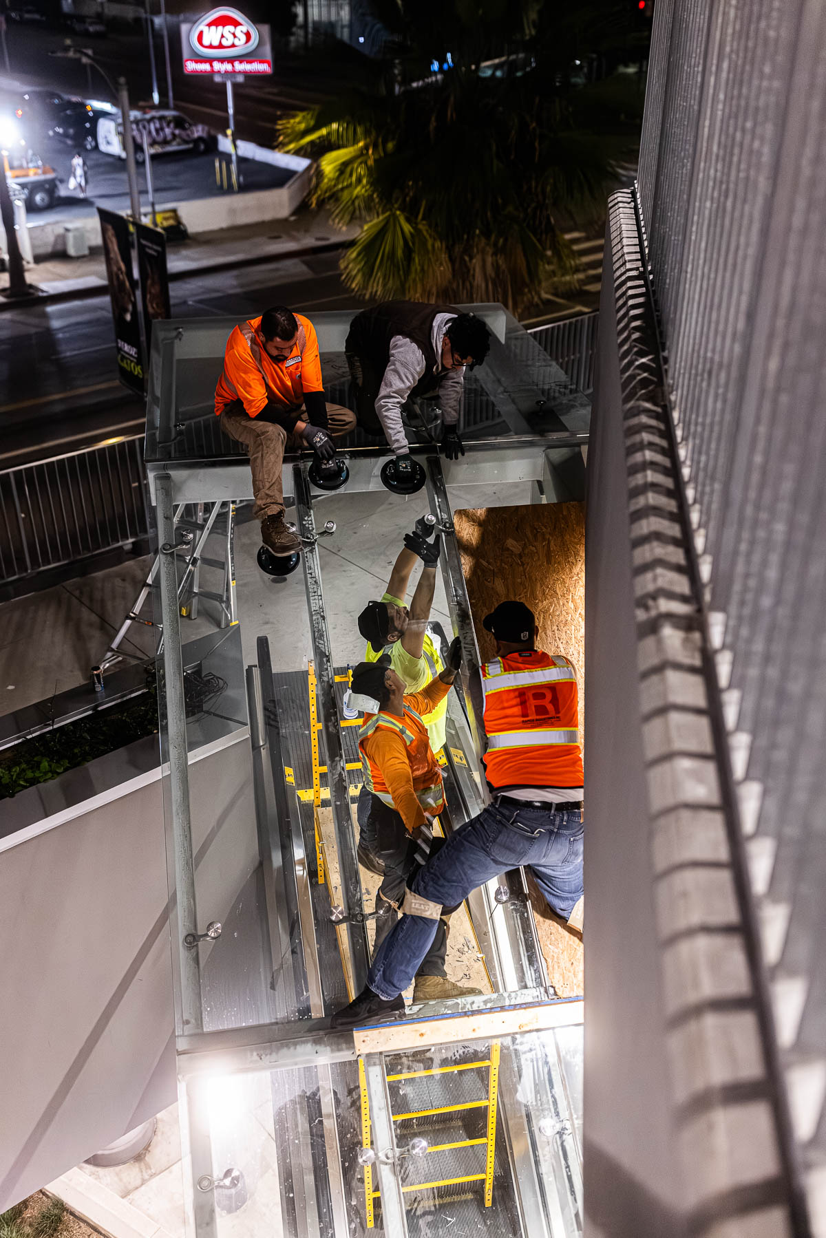 Four construction workers install a large glass panel on an exterior building canopy at night.