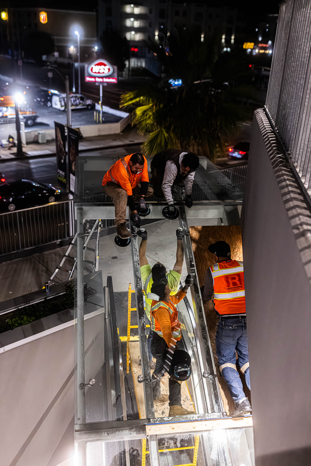 Three construction workers install a large clear glass canopy panel at night at an exterior building entrance.