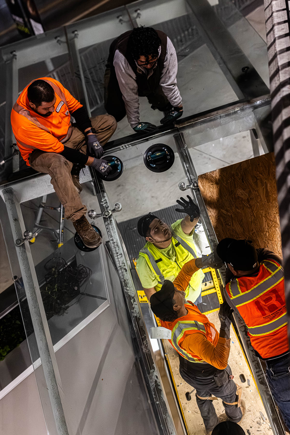 Four glaziers in safety gear work together to install a clear glass canopy panel using suction cups and spider fittings at night.
