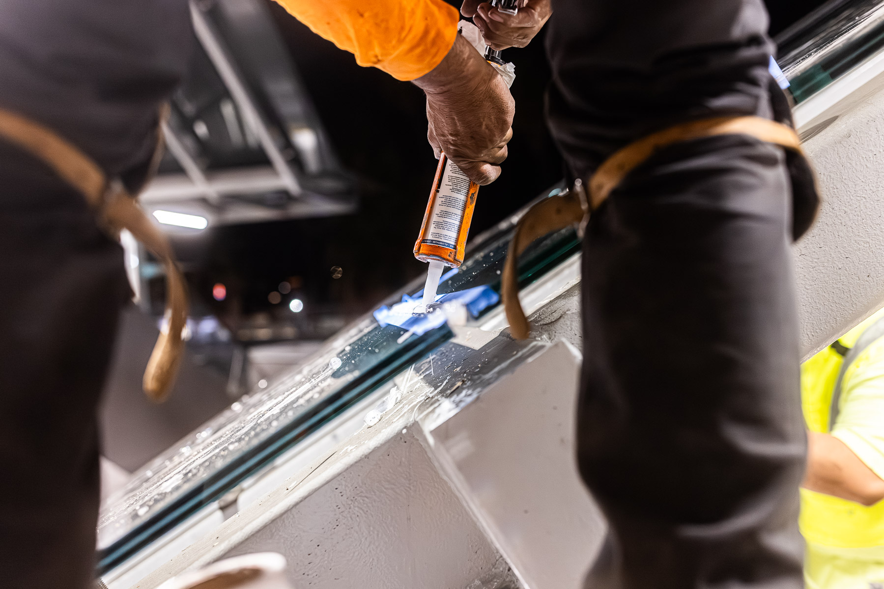 A worker applies sealant to the edge of a clear glass canopy panel at night.