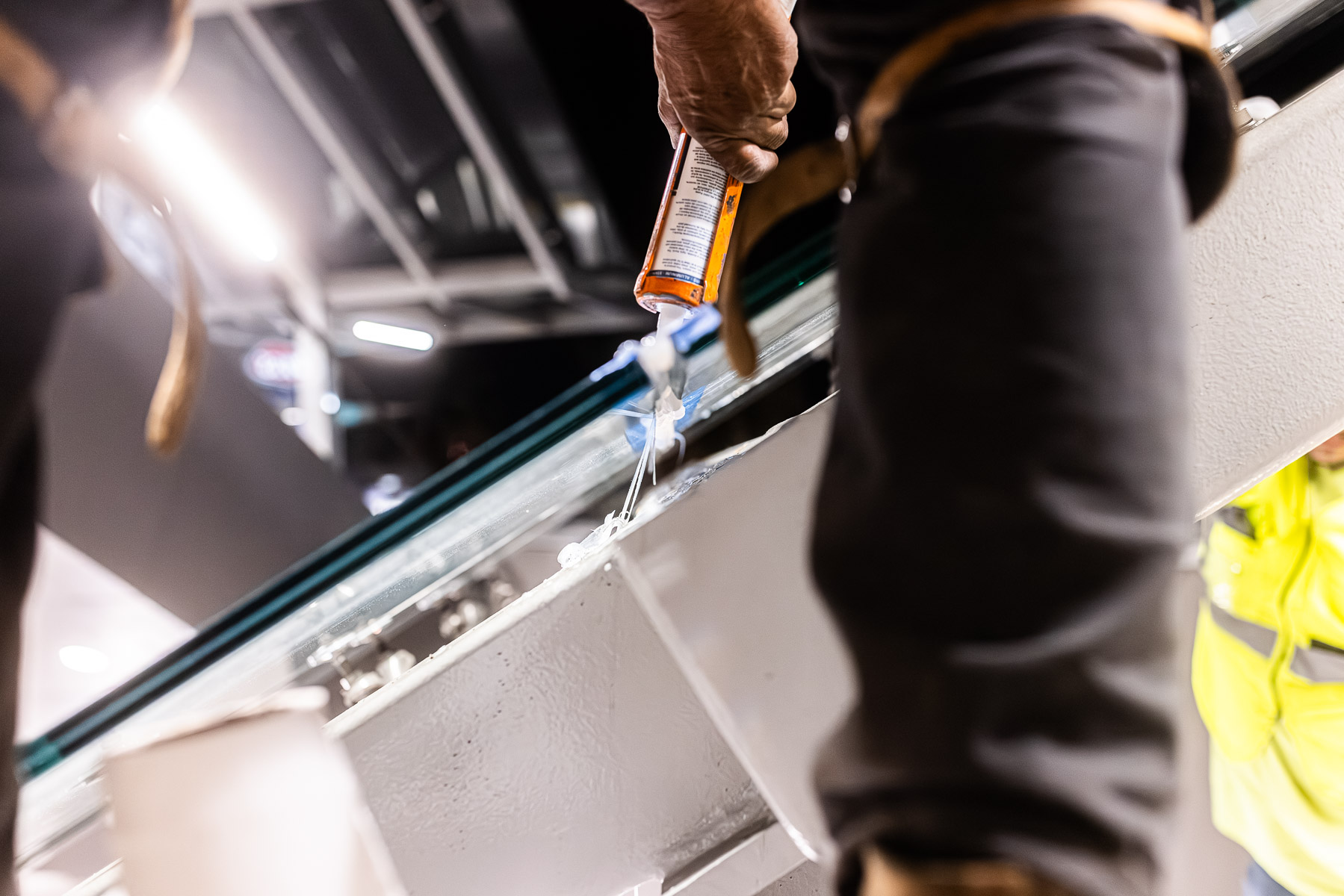 A worker applies sealant to the edge of a clear glass canopy panel installed in a metal frame.
