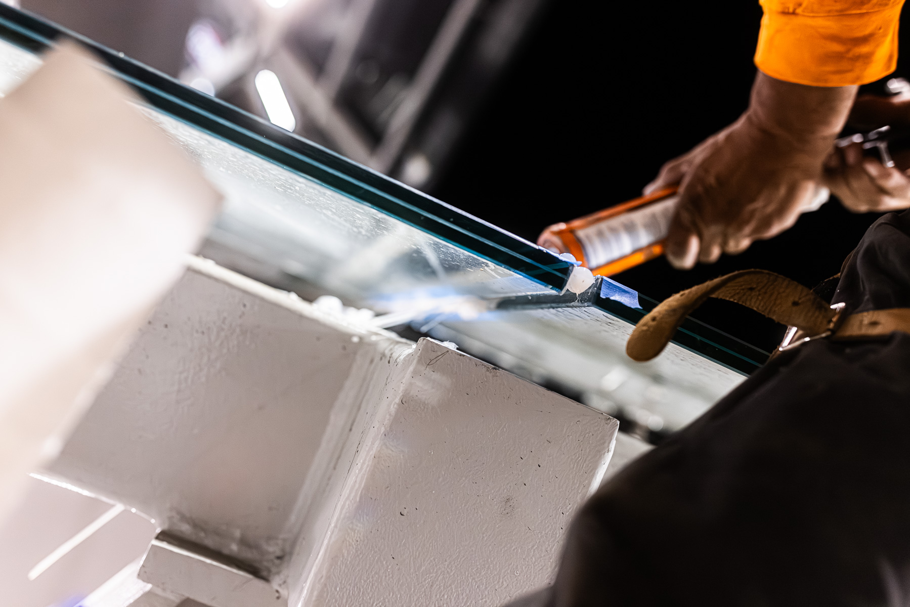 A worker's hands apply sealant to the edge of a clear laminated glass canopy panel.