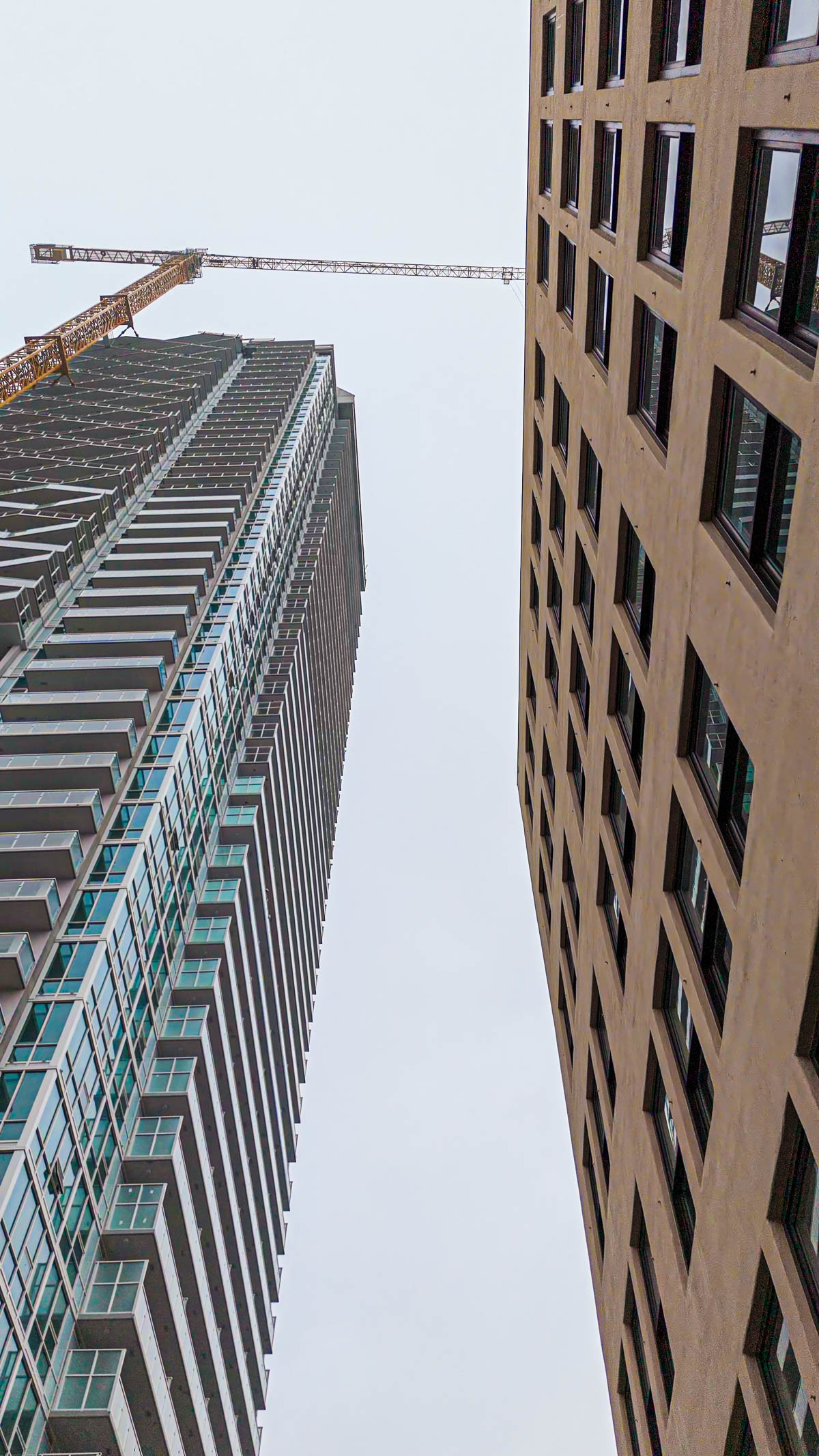 A tall, modern glass building with balconies stands next to a shorter, older building, with a construction crane extending over the modern structure against a grey sky.