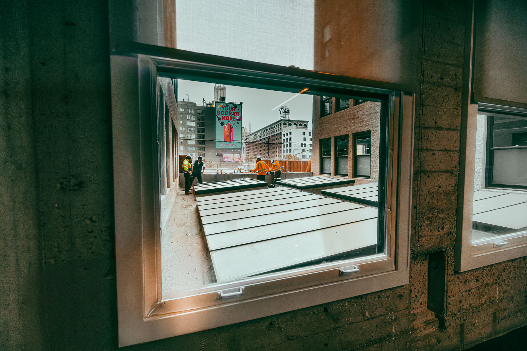 Construction workers on a rooftop are installing large glass panels into an existing skylight frame, viewed through an interior window.