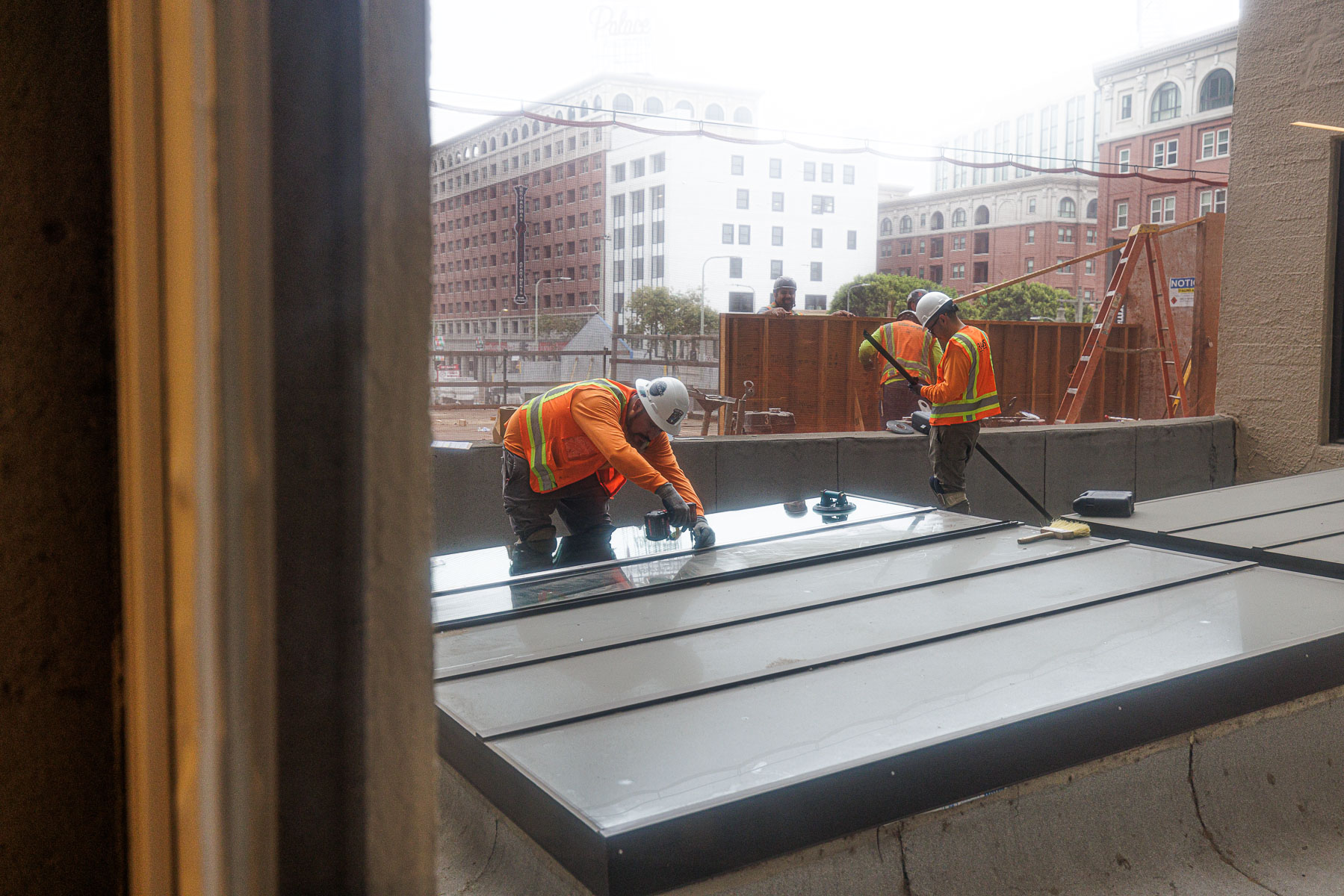 Construction workers in safety gear install a new glass panel into an existing skylight frame on a building rooftop.