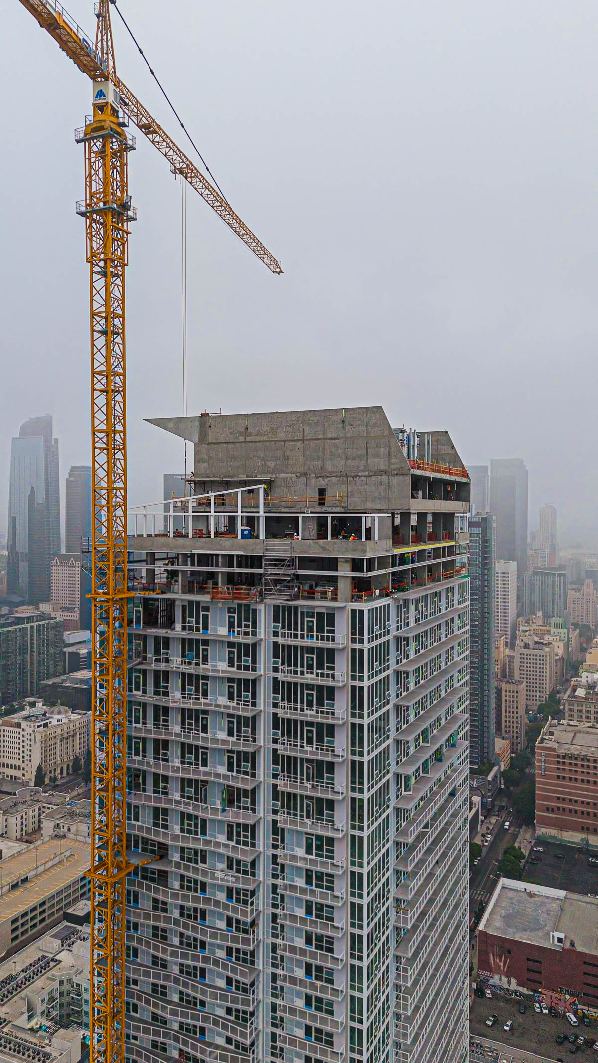 An aerial view of a tall building under construction or renovation in a city, with a large yellow crane beside it under a cloudy sky.