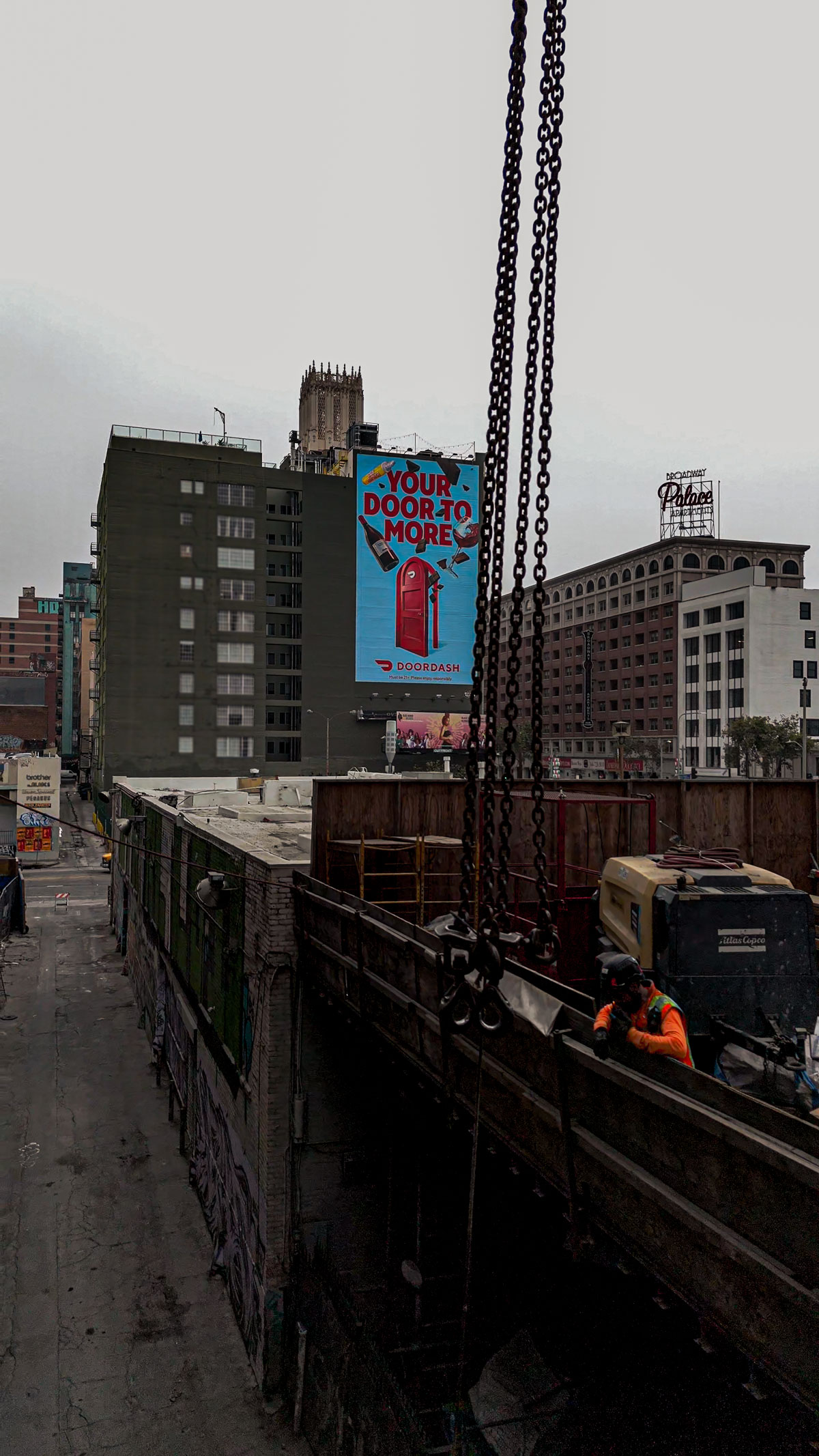 An exterior view of a commercial building in Downtown Los Angeles, showing heavy crane chains overhead and a worker on a construction platform, with city buildings in the background.