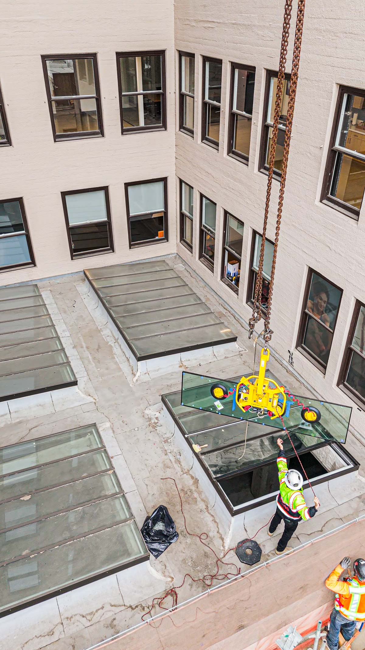 An exterior view shows a worker guiding a large glass panel being lowered by a crane into an open skylight on a building roof.
