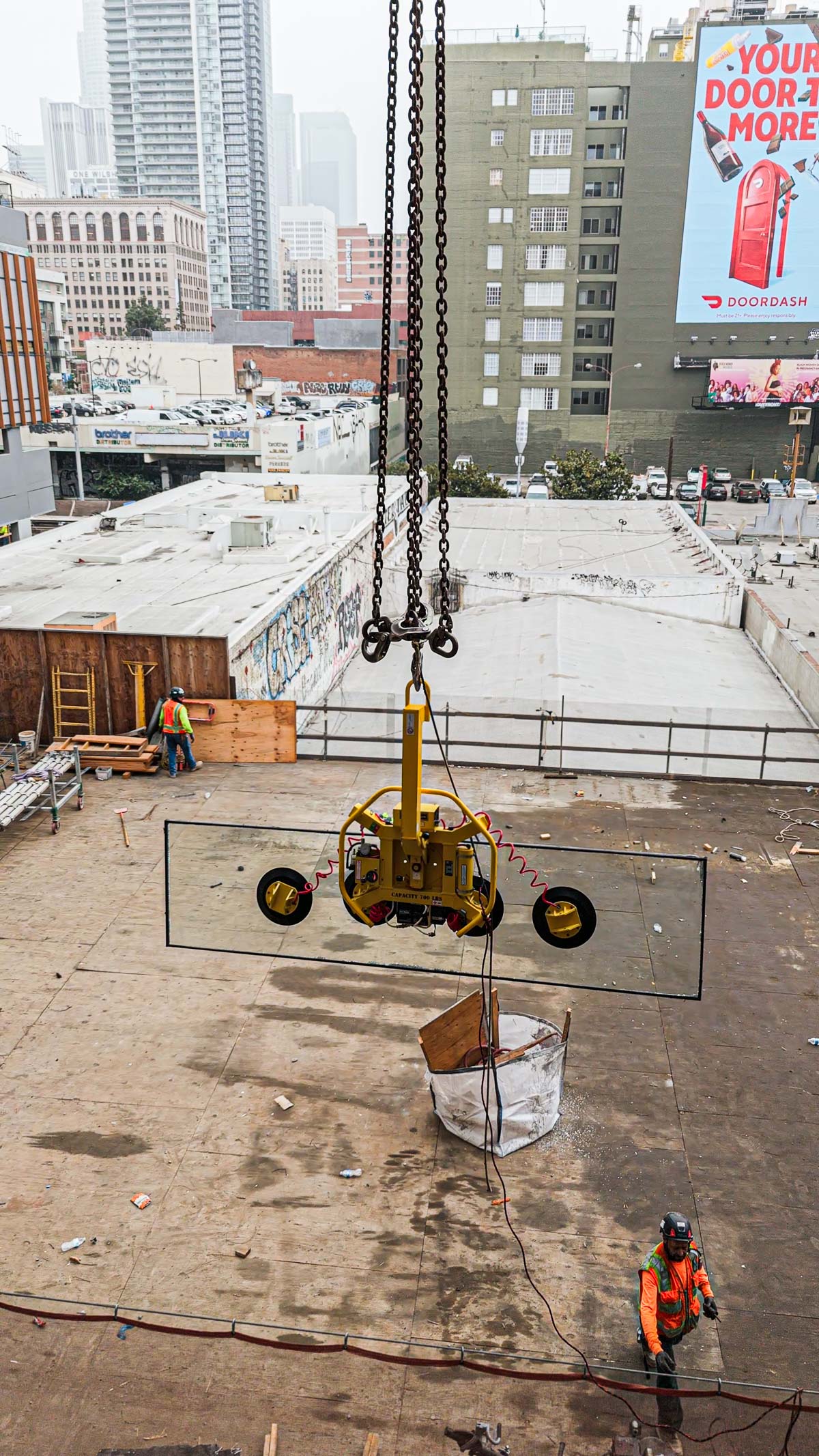A large rectangular glass panel is lowered by a yellow vacuum lifter and crane chains over a construction site in a city.