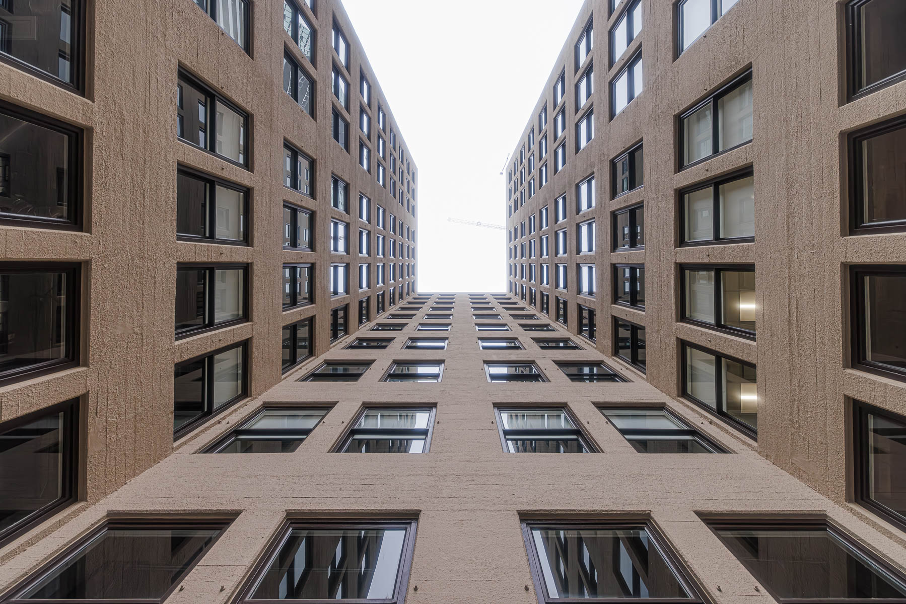 A low-angle view looking up between two tall, light brown commercial buildings with many dark-framed windows, towards a bright sky.