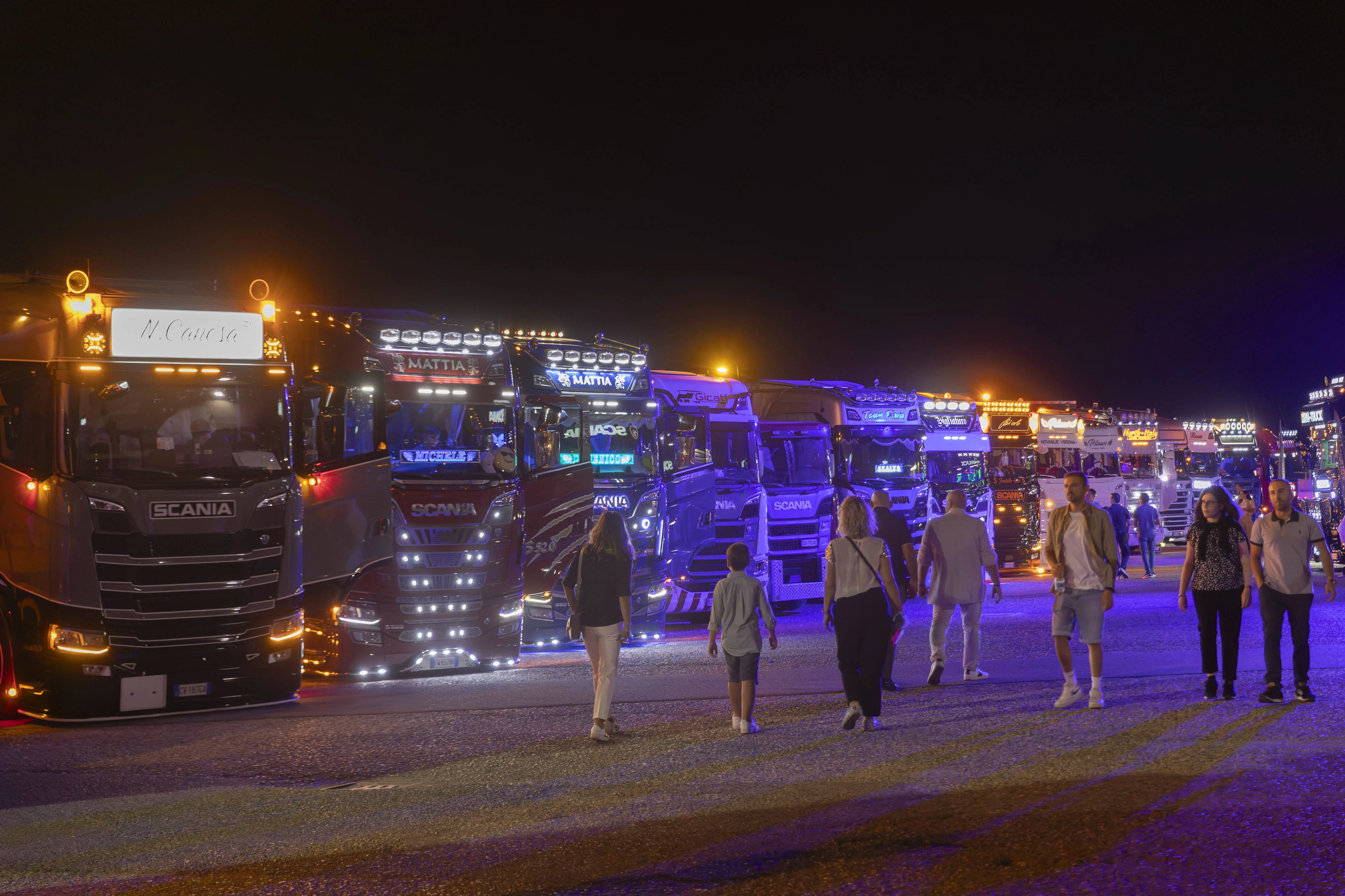 File di camion decorati luminosi a un raduno di camion mentre le persone passeggiano tra loro di notte.