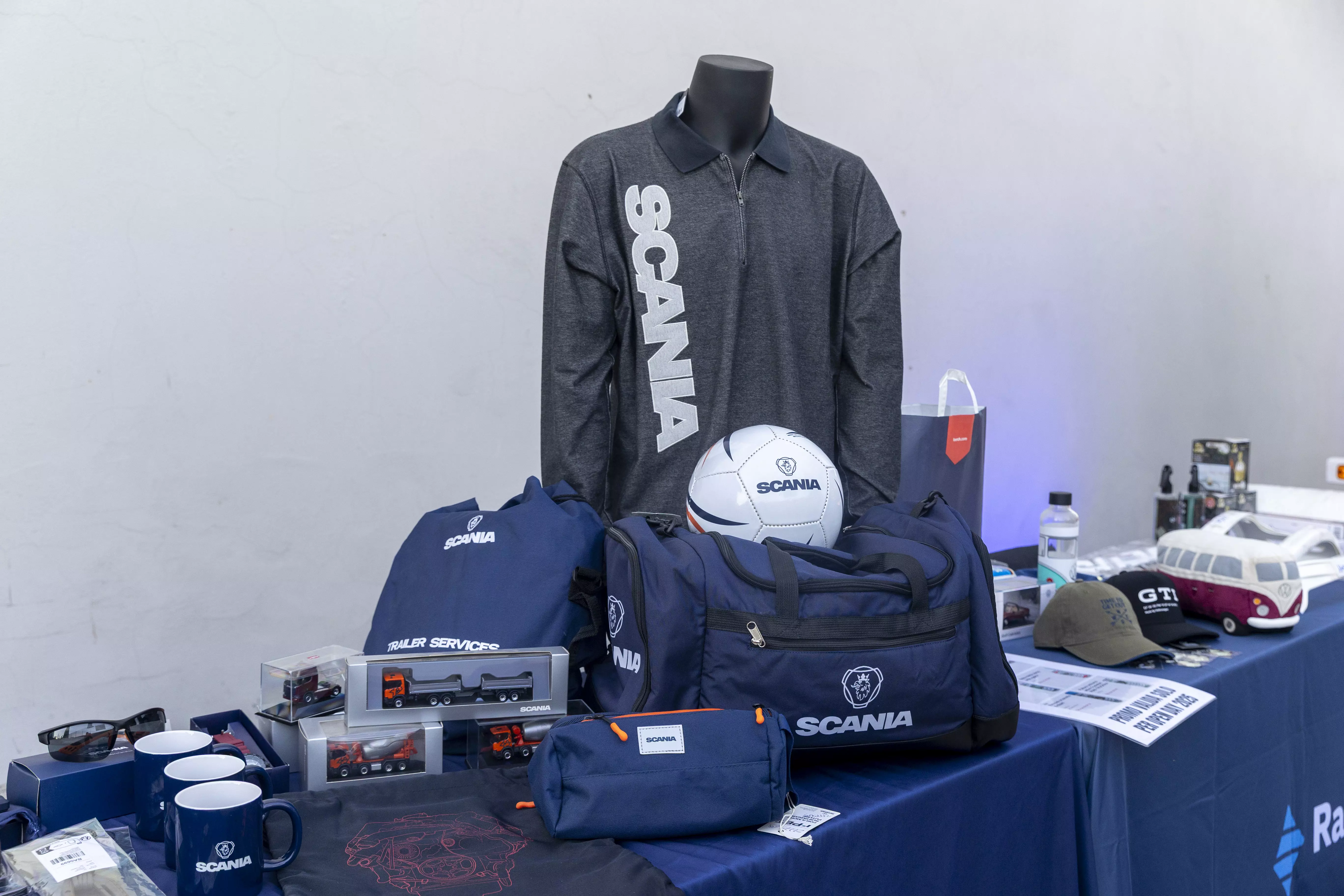 Un tavolo pieno di vari articoli di merchandising Scania, tra cui vestiti, borse e modelli di camion.