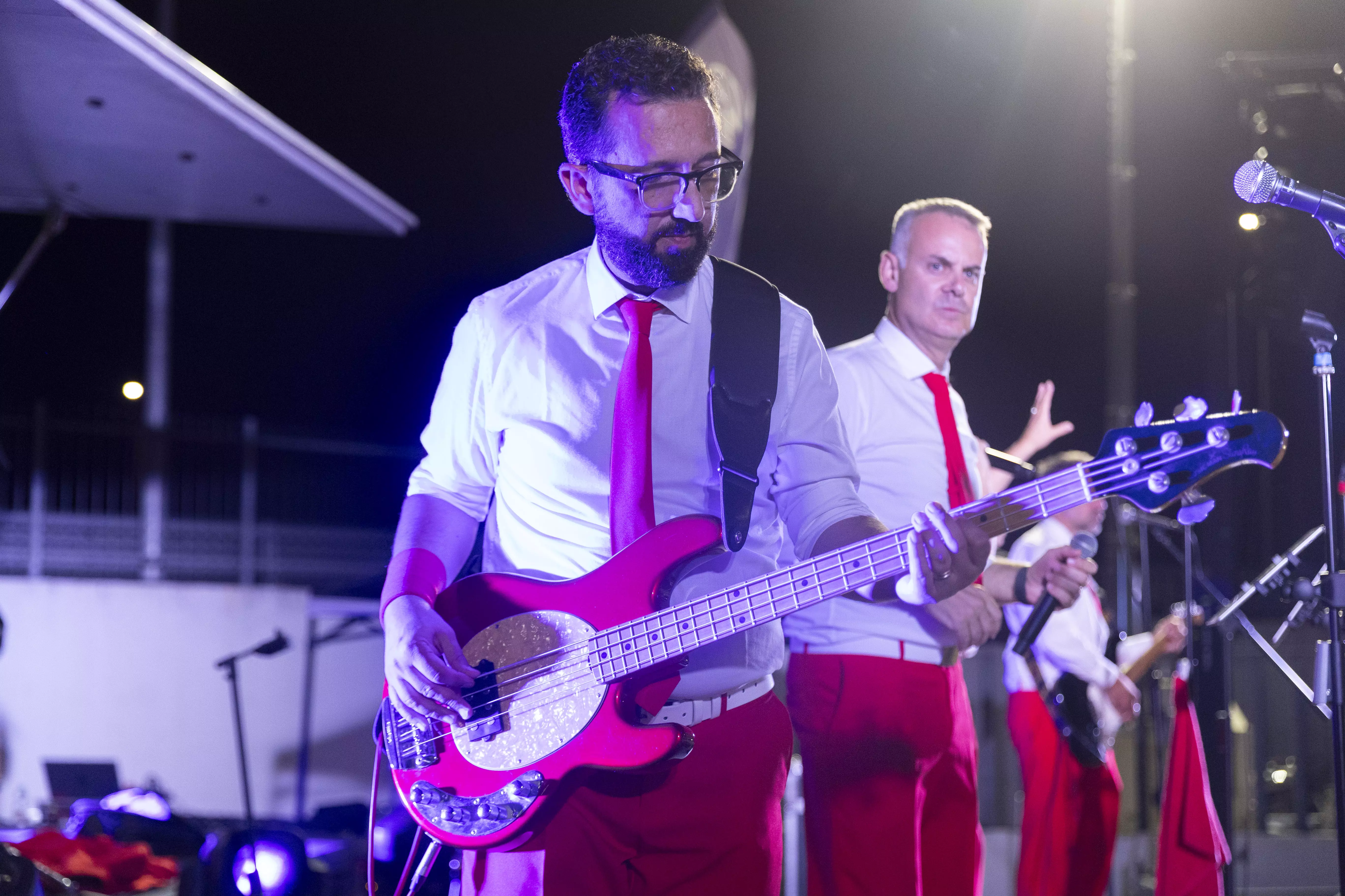 Una band di musicisti si esibisce sul palco con camicie bianche e pantaloni rossi. Il bassista suona un basso rosso.
