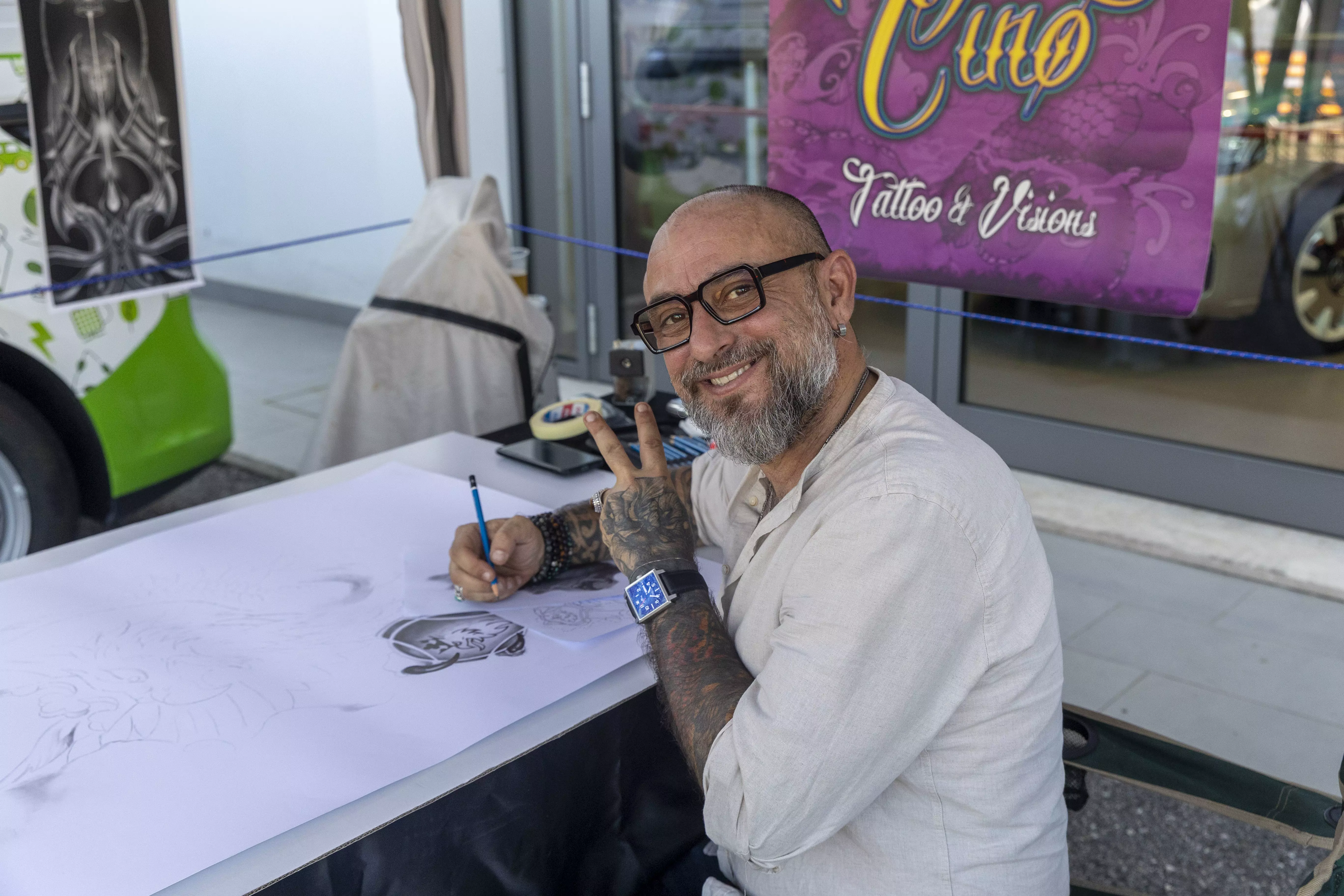 Artista tatuatore sorridente seduto a un tavolo, che fa il segno della pace e disegna.