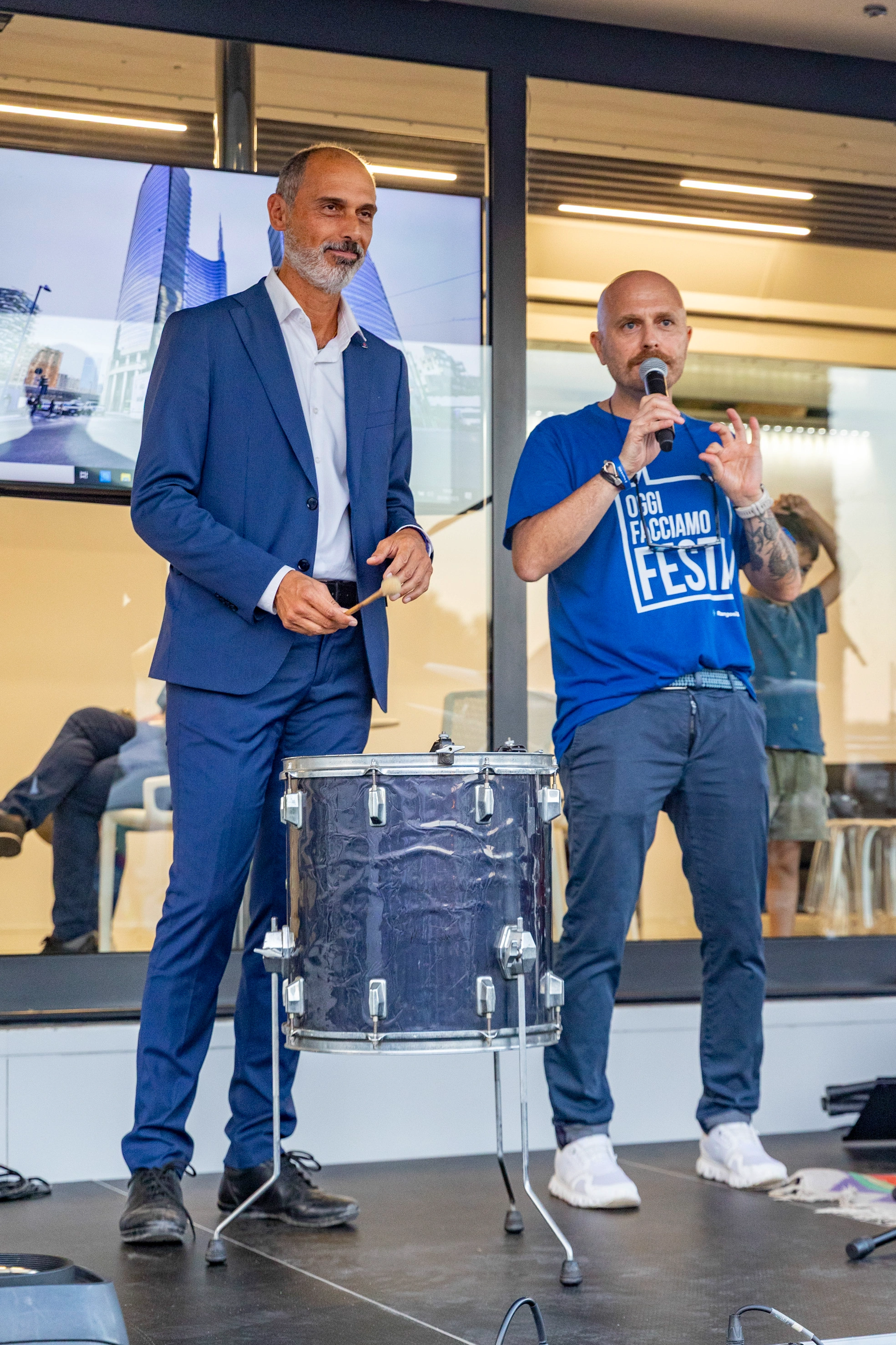 Due uomini su un palco, uno con un abito blu e l'altro con una maglietta blu e un microfono, con una batteria in mezzo a loro.