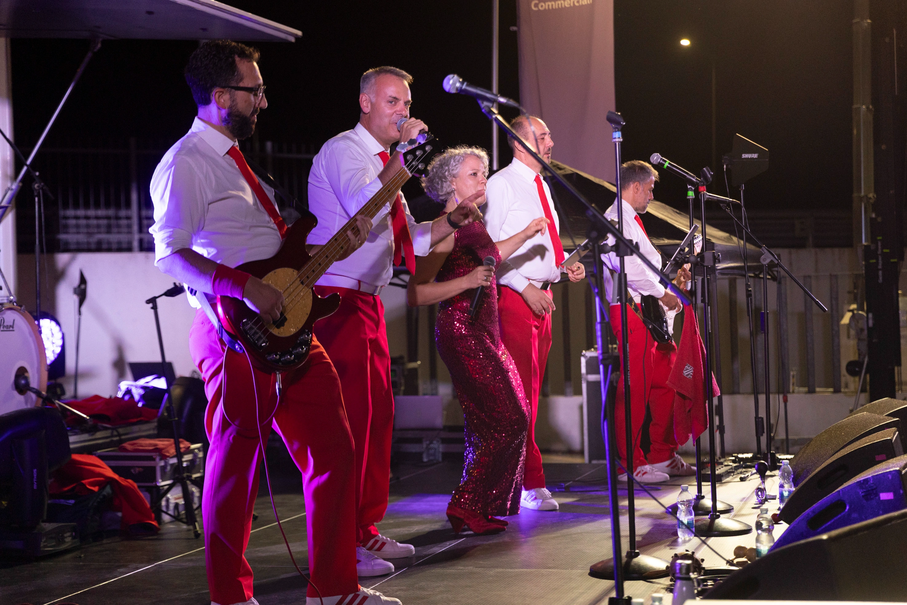 Una band di musicisti si esibisce sul palco indossando camicie bianche e pantaloni rossi con una cantante in abito rosso.