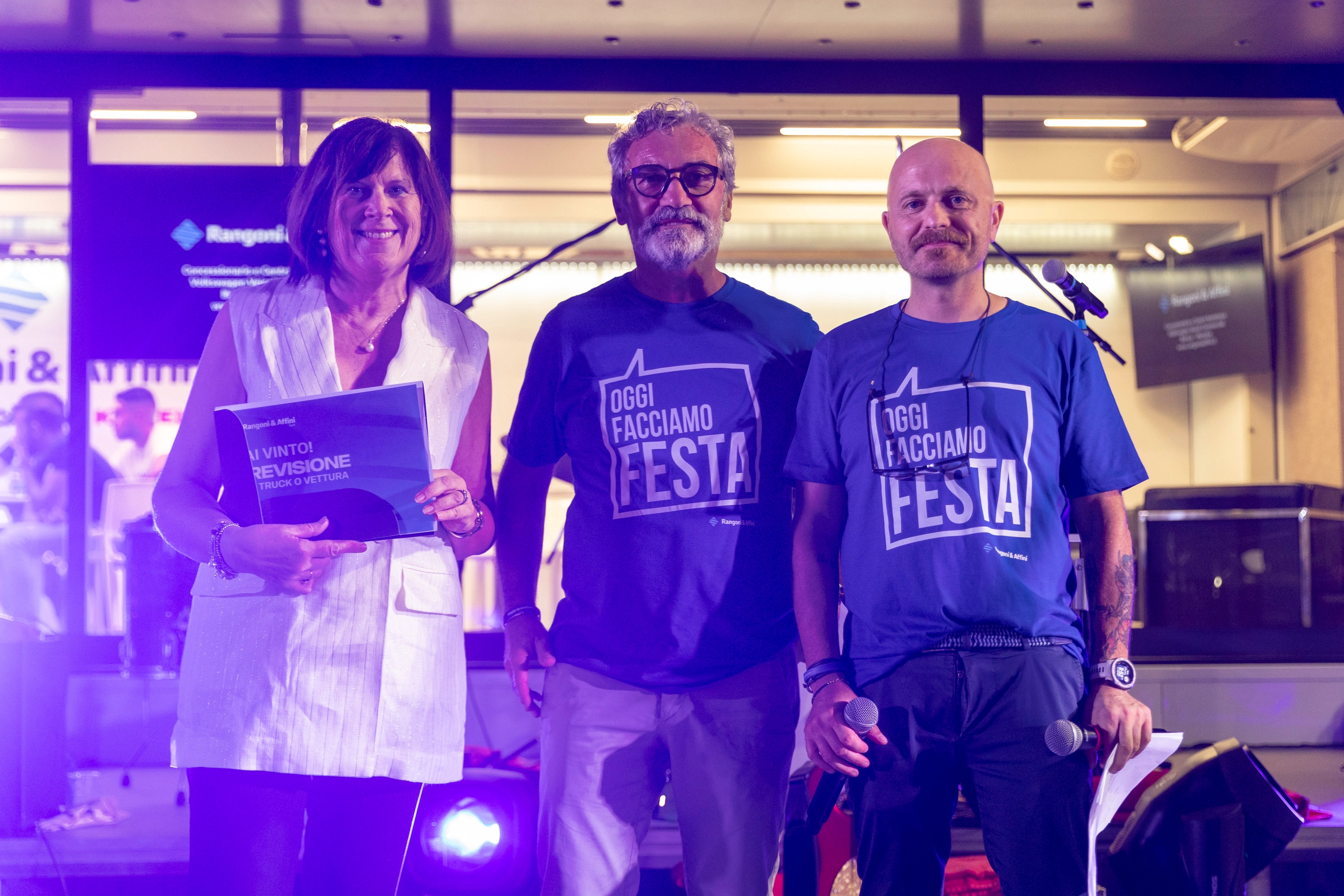Tre persone sorridenti sono in piedi su un palco con i loghi di Rangoni & Affini. La donna tiene un cartello con la scritta "Hai vinto! Revisione."