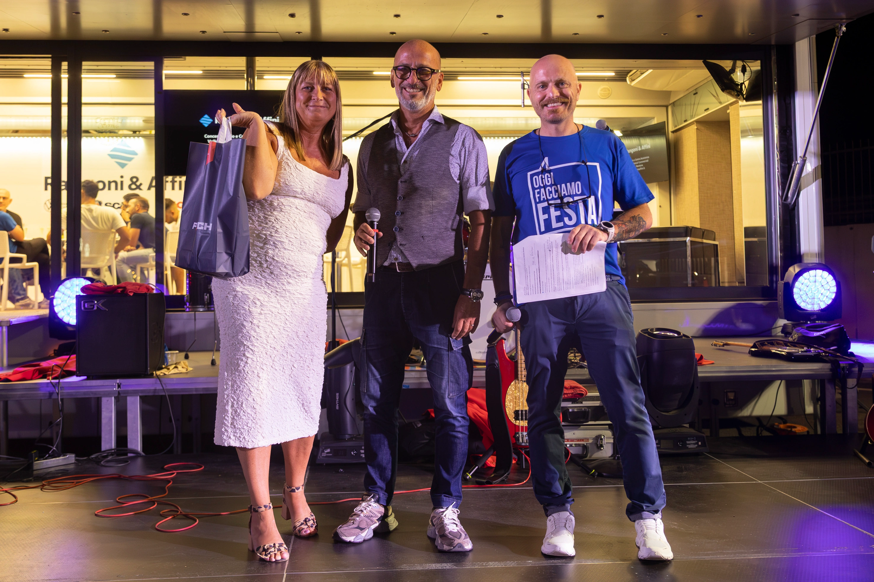 Tre persone sorridenti sul palco con microfoni, una donna con una borsa.