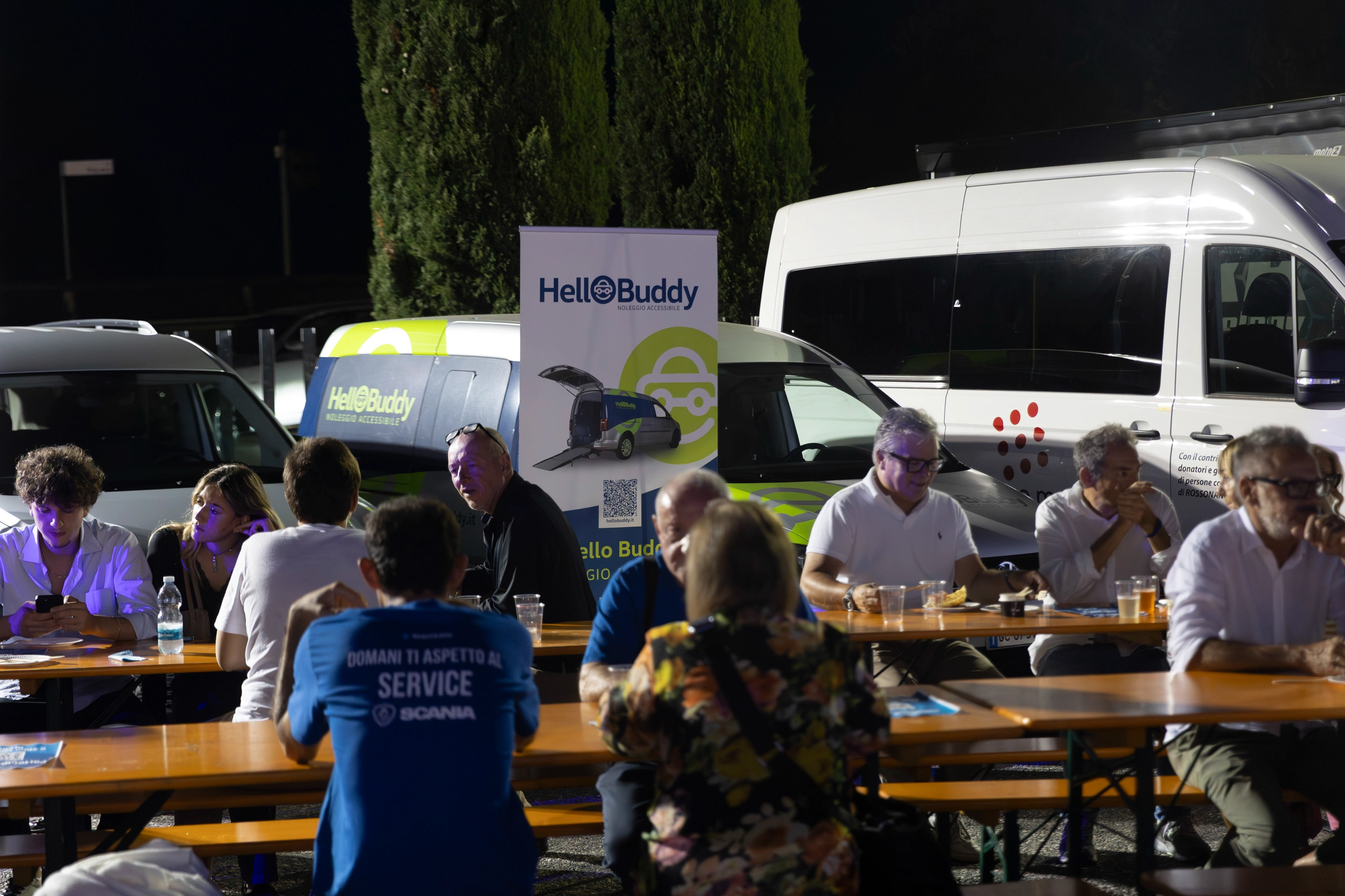 Un gruppo di persone sedute a tavoli di legno davanti a furgoni Hello Buddy durante un evento notturno.