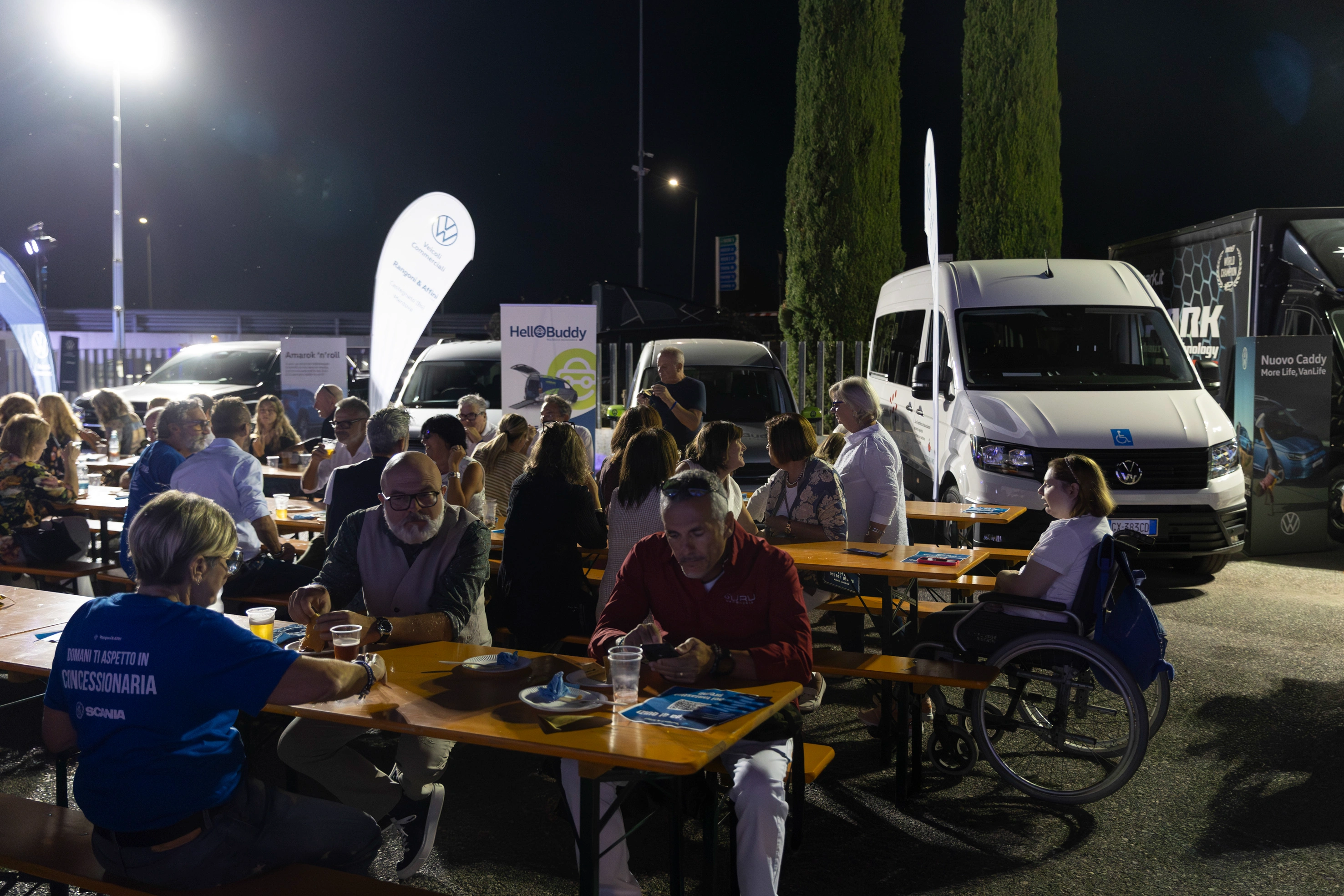Una folla si riunisce all'aperto con tavoli e furgoni Volkswagen sullo sfondo.