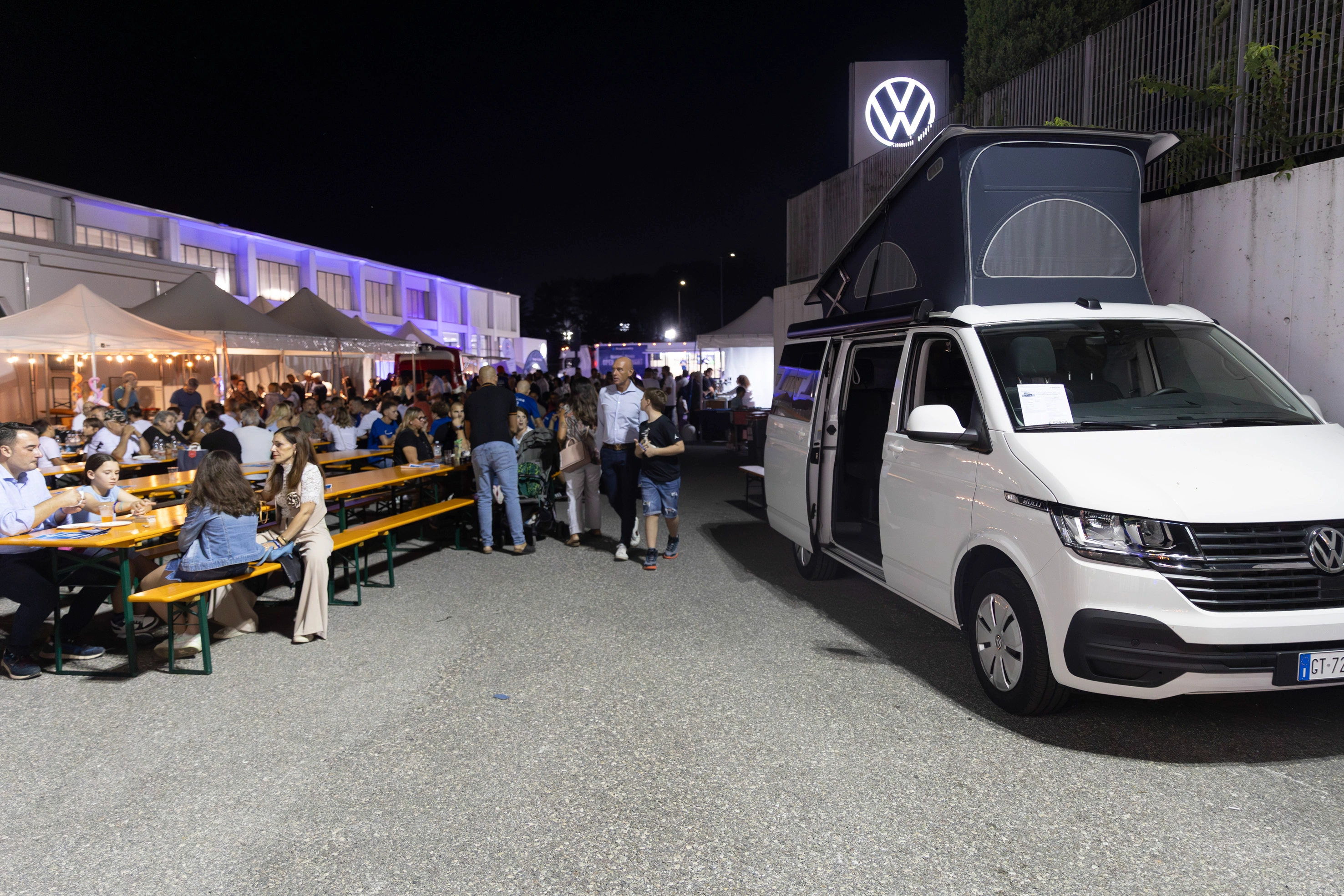 Gente riunita intorno a tavoli di legno con furgone Volkswagen in mostra di sera.