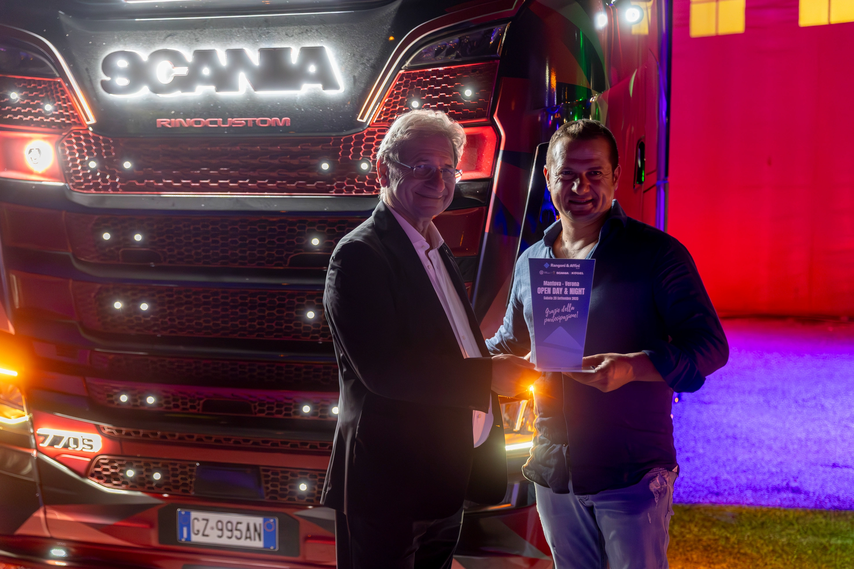 Due uomini posano di fronte a un camion Scania con una targa con la scritta "GZ-995AN", tenendo in mano un trofeo.