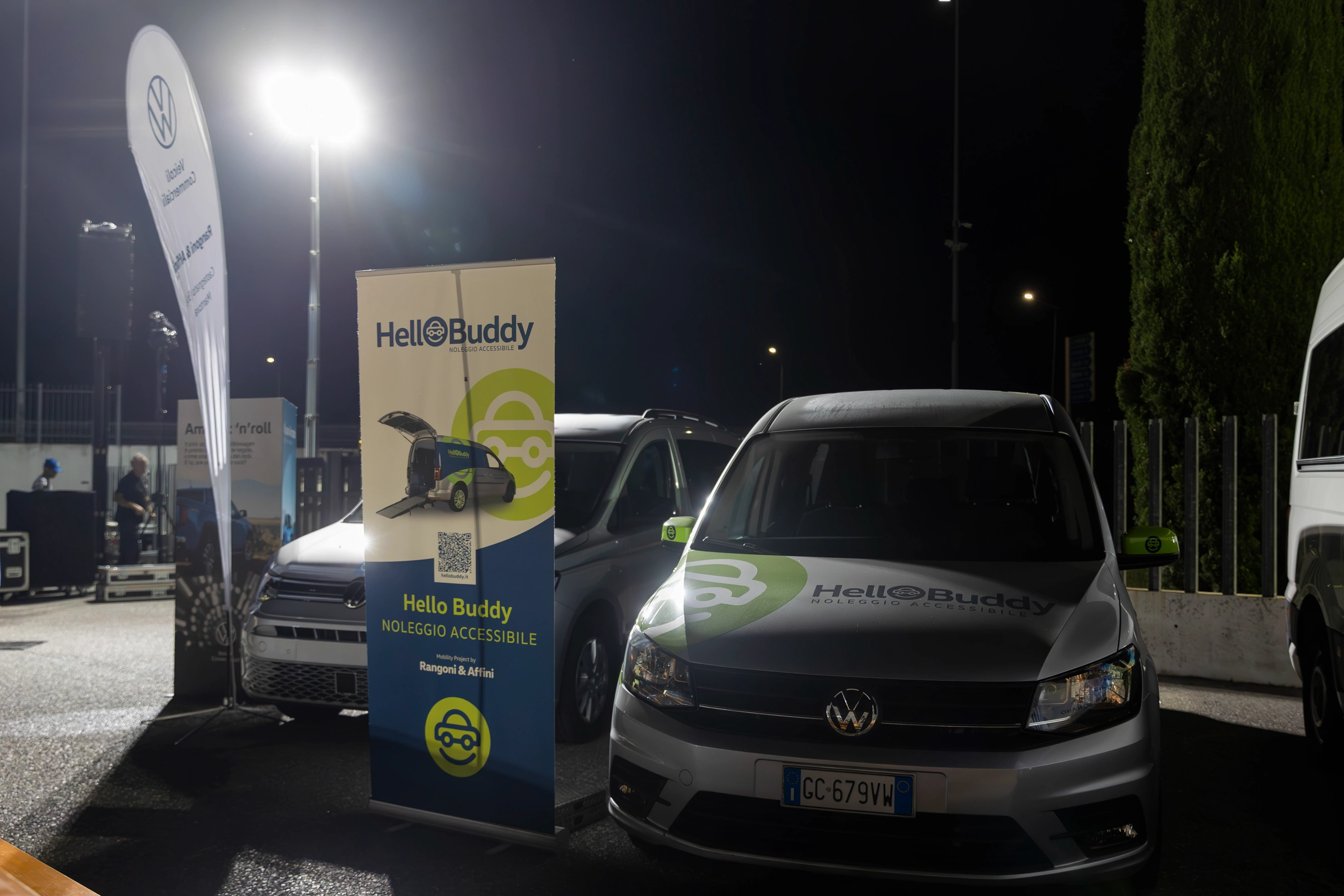 Auto Hello Buddy con grafica verde, parcheggiata di notte accanto a striscioni con logo.
