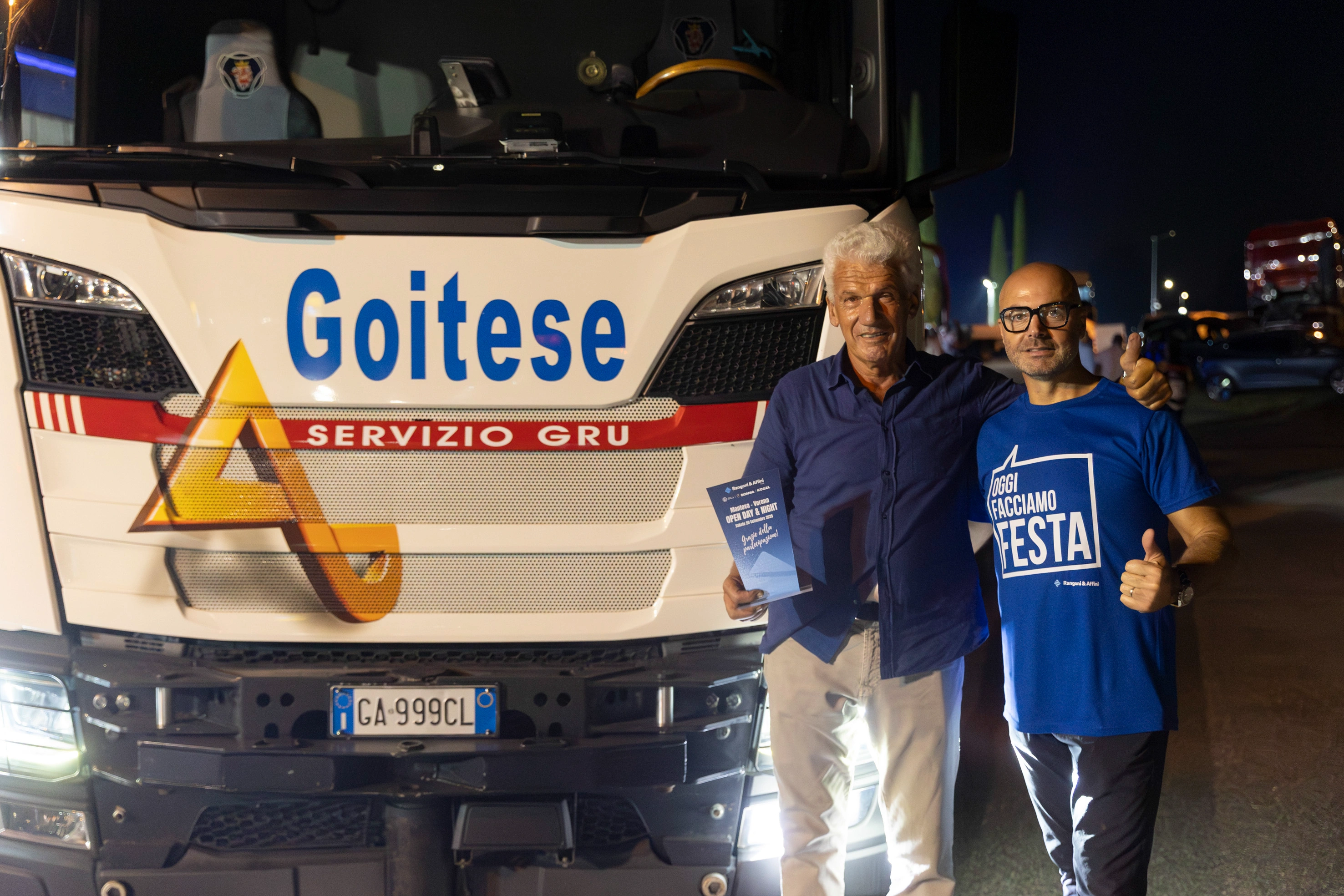 Due uomini in posa davanti a un camion Goitese con il logo Servizio Gru, che tiene in mano un opuscolo e indossa una maglietta blu con la scritta "Oggi Facciamo Festa".