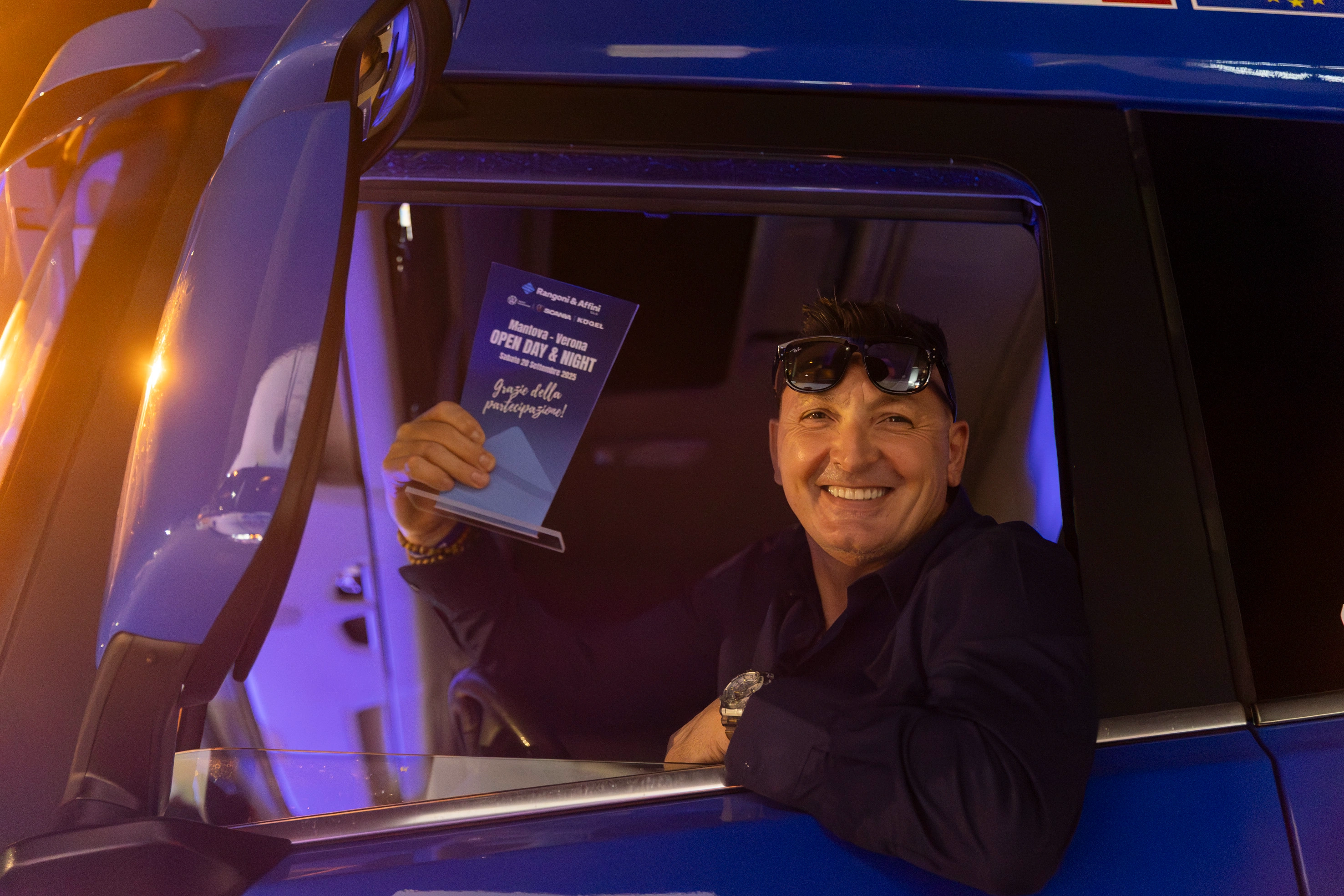 Un uomo sorridente sporge la testa dal finestrino di un camion e mostra un volantino dell'evento.